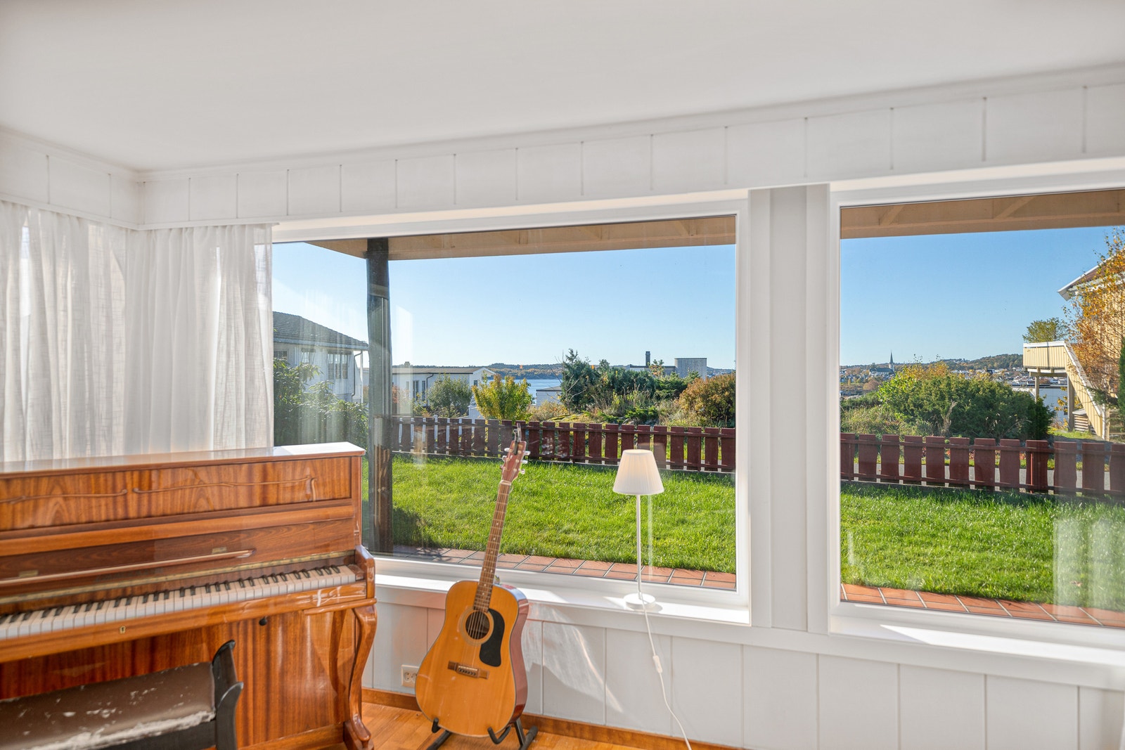 Pianokrok med gitar og store vinduer mot hage og sjø. Galleribilde