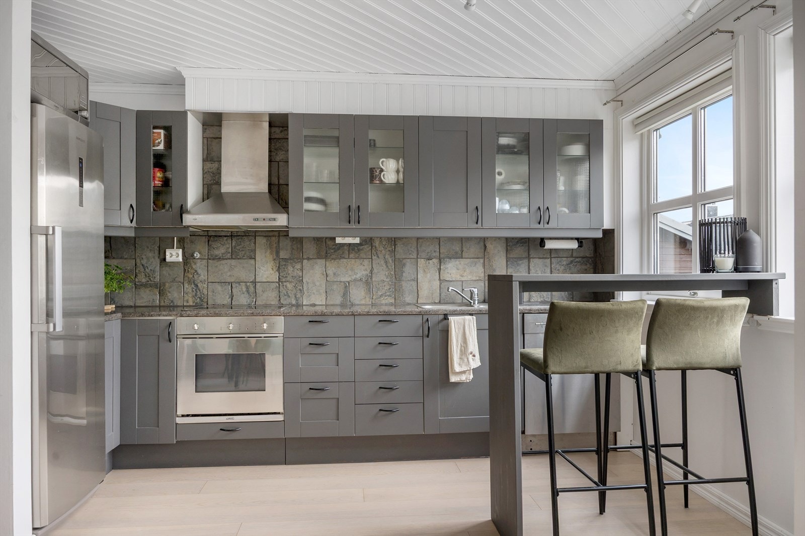 Fliser mellom benkeplaten og overskapene gir et stilrent uttrykk, og kjøkkenet er utstyrt med integrert steketopp og koketopp. Oppvaskmaskin og kjøleskap ikke er integrert, men begge medfølger handelen. Galleribilde