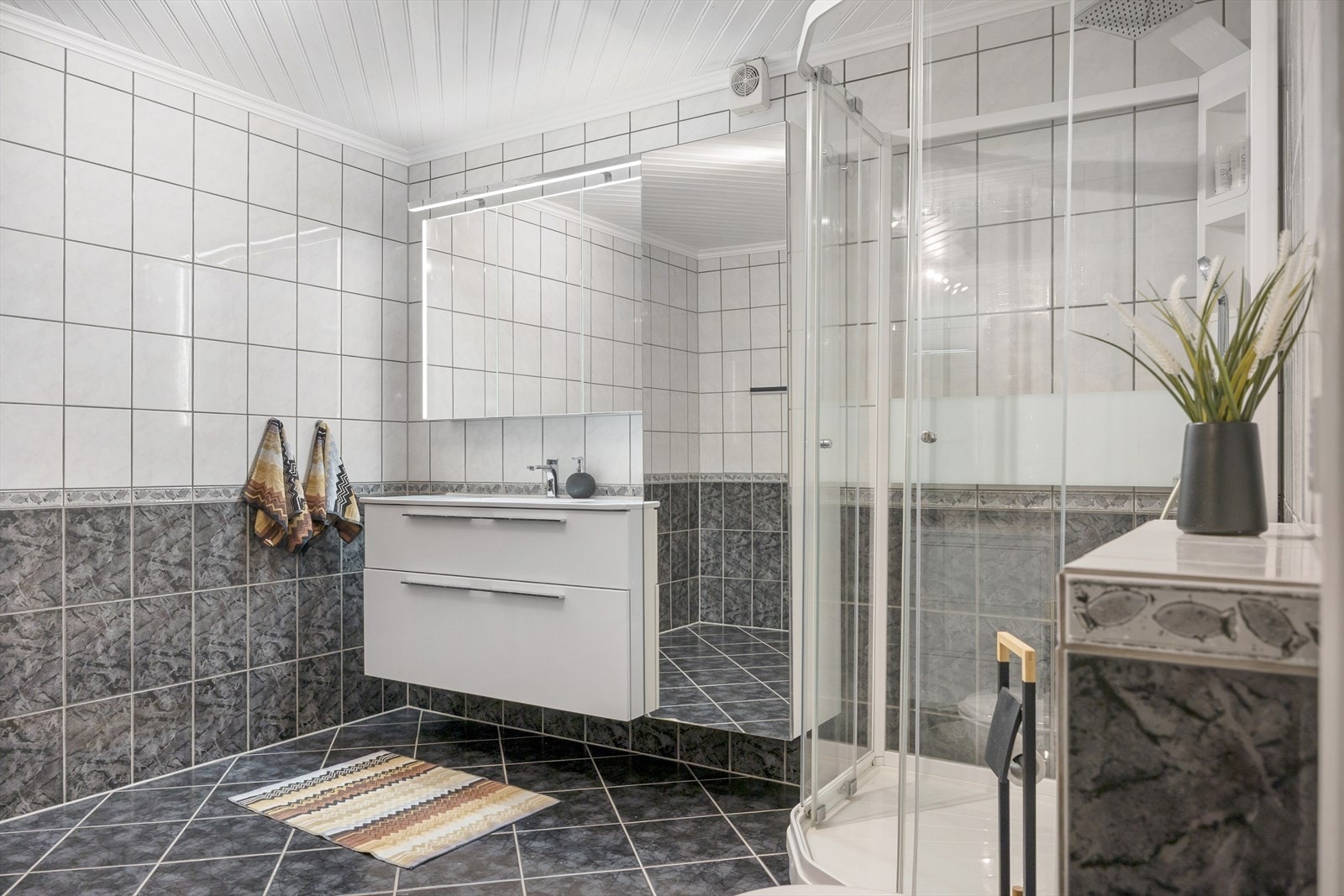 Badet er romslig med helflisede vegger og varmekabler i gulvet. Innredningen består av en servant nedfelt i benkeskap, speilskap med lys, vegghengt toalett og dusjkabinett. Her er det tilrettelagt for høy komfort i hverdagen. Galleribilde