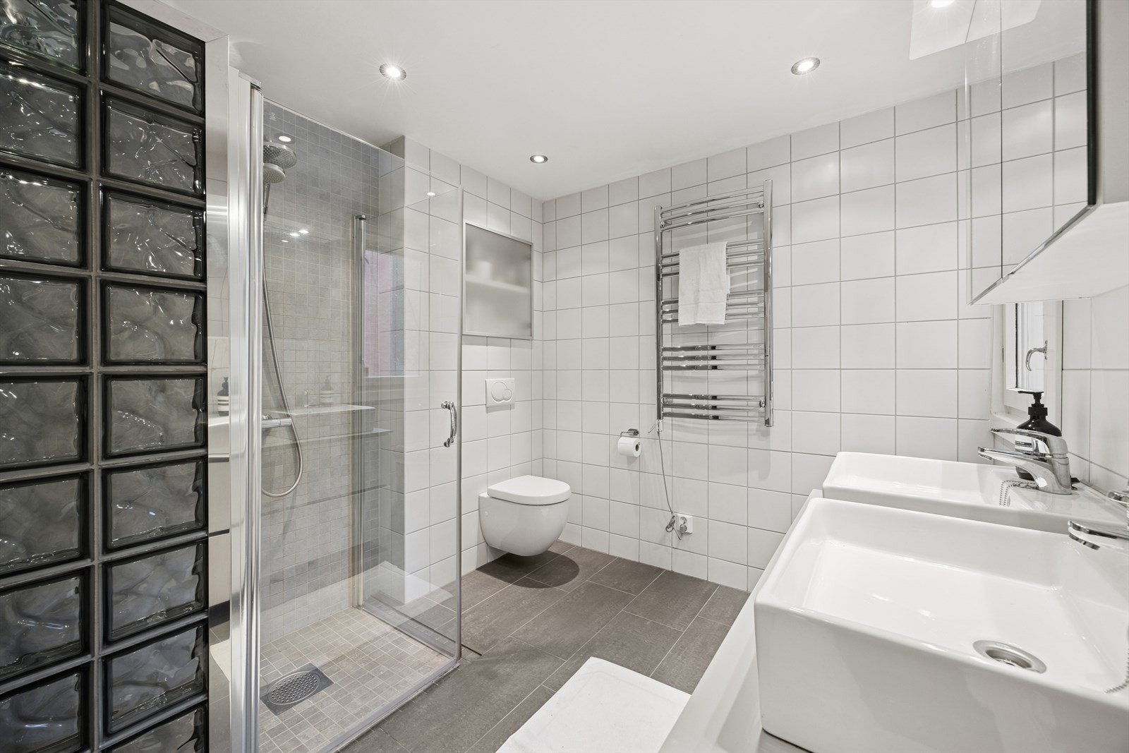 Badet er innredet med et vegghengt klosett, servant nedfelt i innredning, speilskap med lys og et dusjhjørne med svingbare glassdører. Rommet har ventilasjon via en elektrisk styrt vifte. Galleribilde