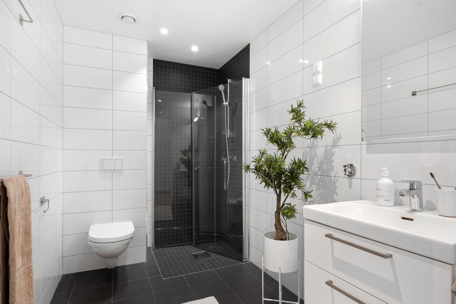 Badet ligger praktisk plassert i tilknytning til entréen og fremstår delikat med hvite veggfliser og mørke gulvfliser. Innredningen består av nedfelt servant med speil over, veggmontert toalett og dusjhjørne med mosaikkfliser og innfellbare glassdører. Galleribilde