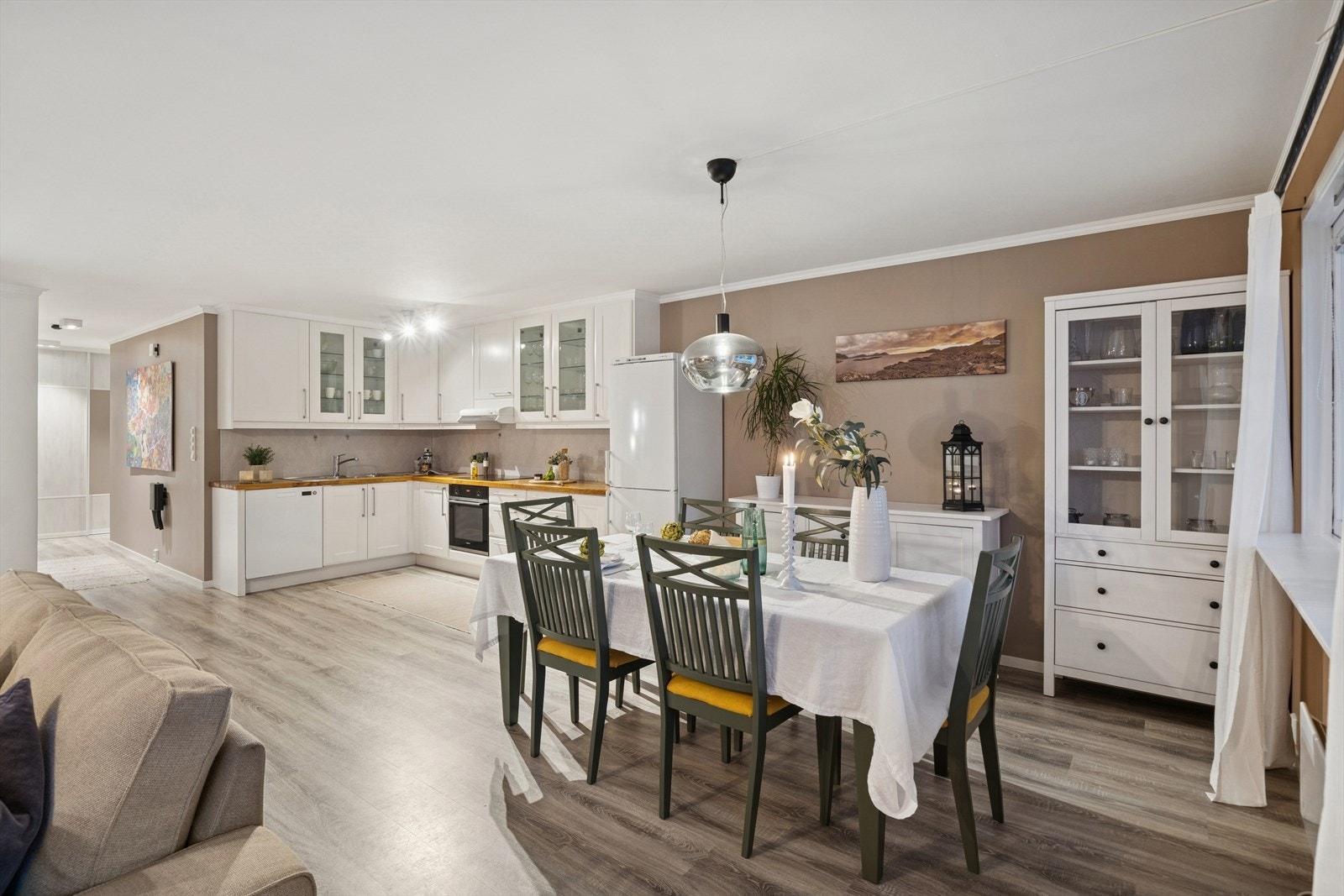 Rommet har en åpen og innbydende løsning som kombinerer kjøkken og spisestue på en harmonisk måte. Laminatgulvet i naturtro trestruktur binder sonene sammen og skaper en lun helhet. Et perfekt rom for både hverdagsmåltider og sosiale sammenkomster. Galleribilde