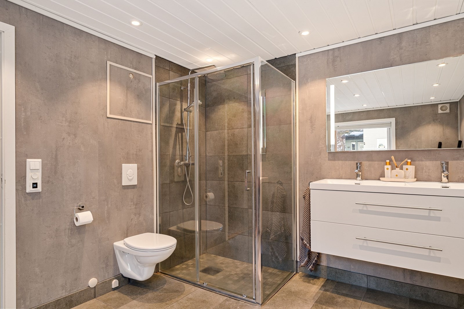 Rommet er utstyrt med flislagt gulv med varmekabler, downlights i tak og en romslig dusjsone med waterfall-dusj. Videre har badet vegghengt toalett og dobbel servant, noe som gir både god funksjonalitet og et stilrent uttrykk. Galleribilde