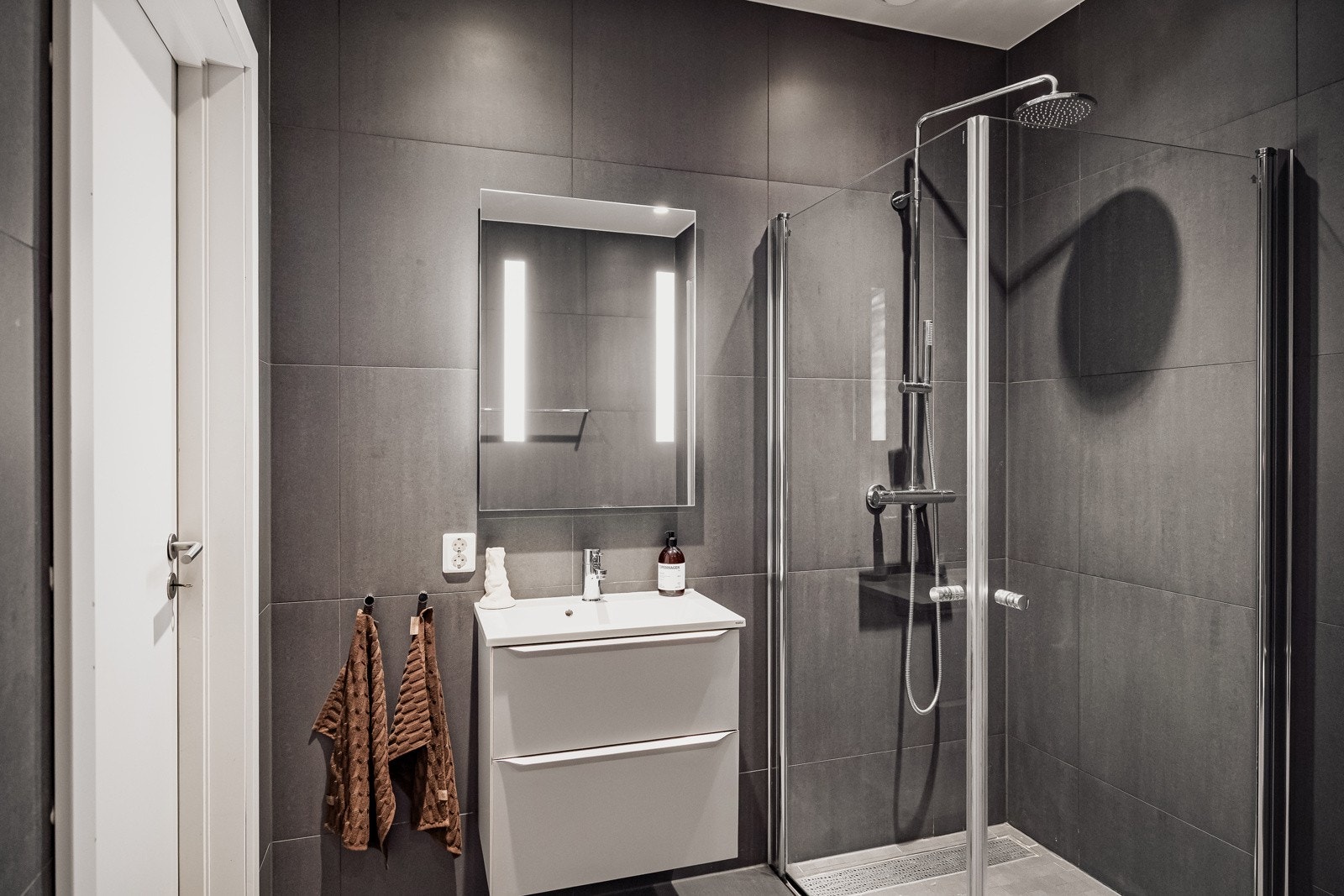 Moderne baderomsinnredning med skuffer og heldekkende servant, speil med integrert belysning over. Baderommet er også utstyrt med dusjhjørne med innfellbare glassdører og både hånd- og regnfallsdusj, samt vegghengt toalett. Galleribilde