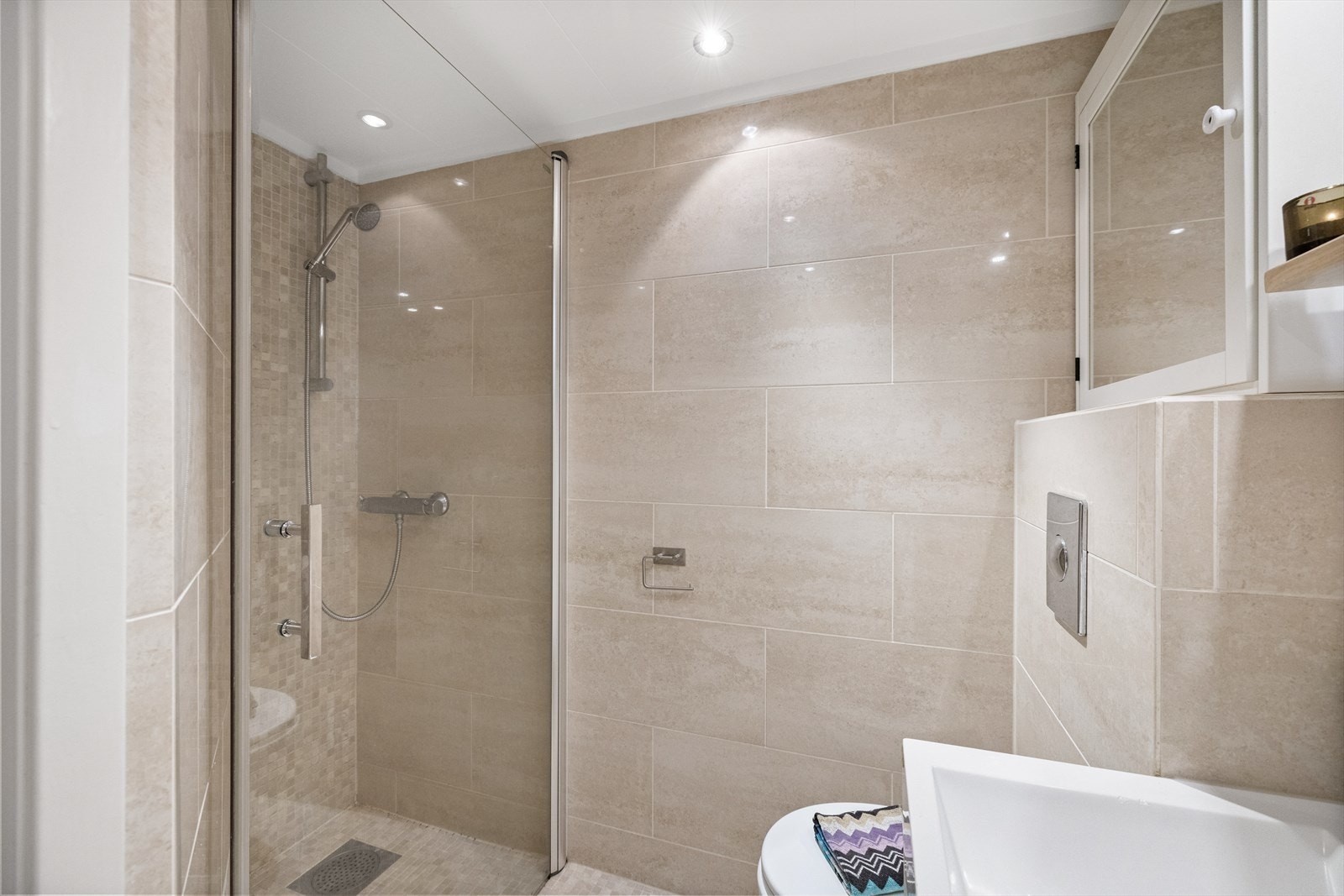 Badet er innredet med vegghengt toalett, servant med praktisk innredning og dusjhjørne med glassvegger. Galleribilde