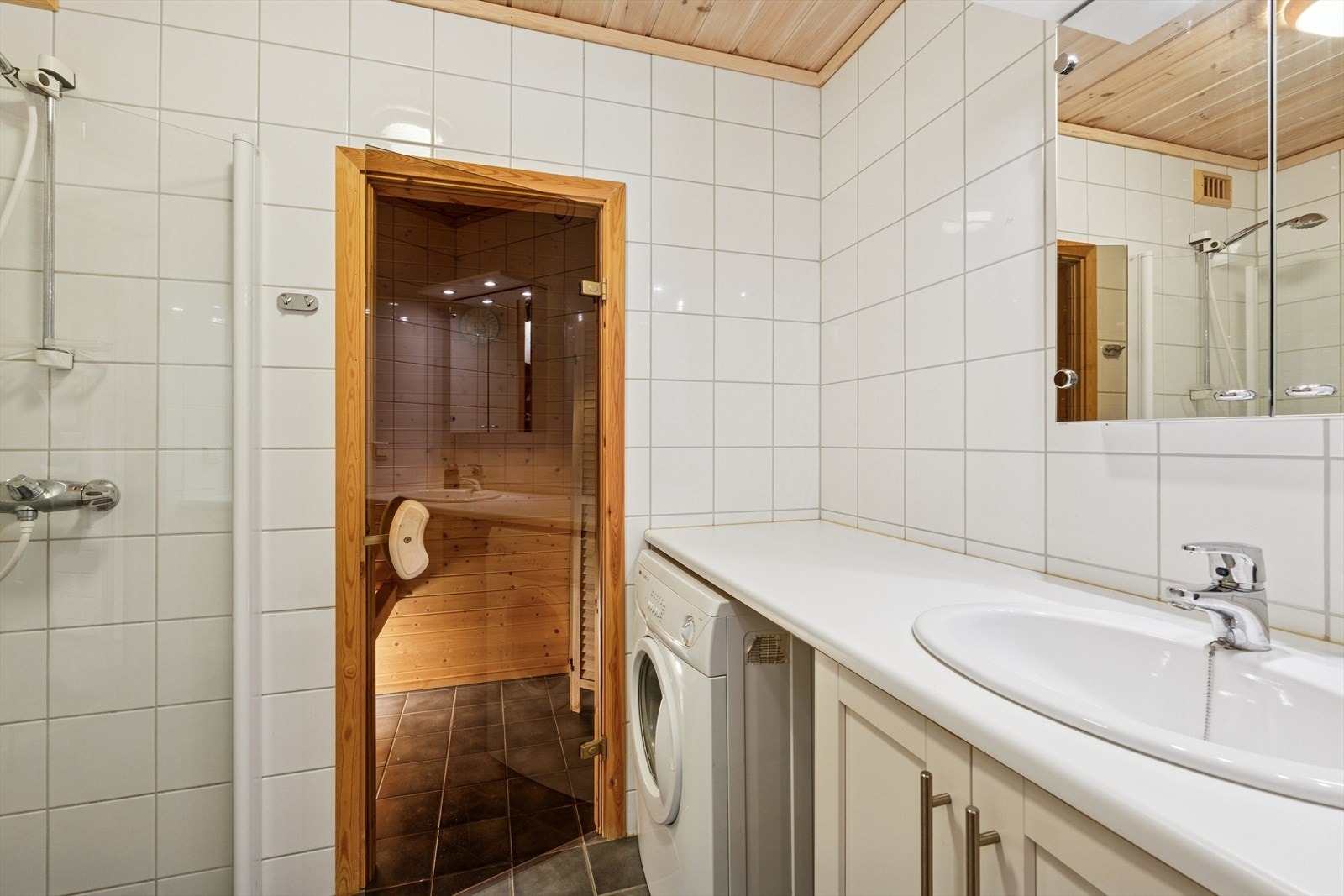 Baderommet er innredet med benk med vaskemaskin under, baderomsinnredning, nedfelt servant, veggmontert klosett og dusj med innfellbare glassvegger. Galleribilde