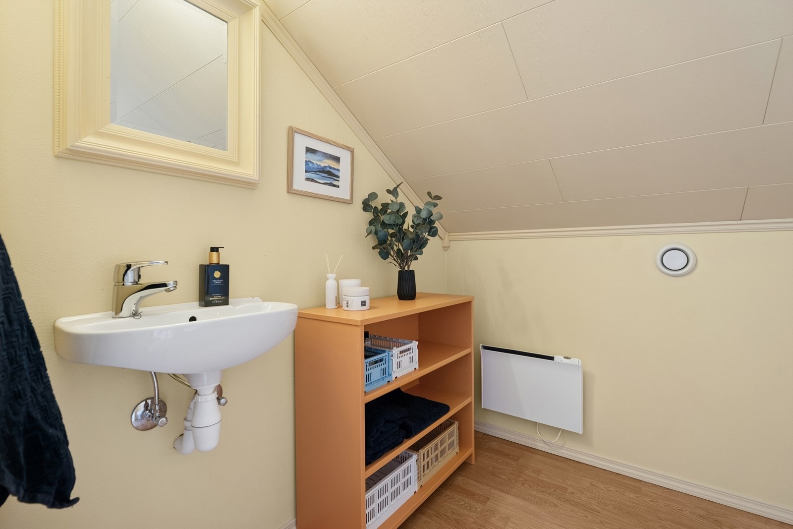 Toalettrommet har laminatgulv, vegger med malt tapet og folierte trefiberplater i himlingen. Rommet er innredet med en enkel servant og et vegghengt toalett, som gir et ryddig og moderne uttrykk. Galleribilde