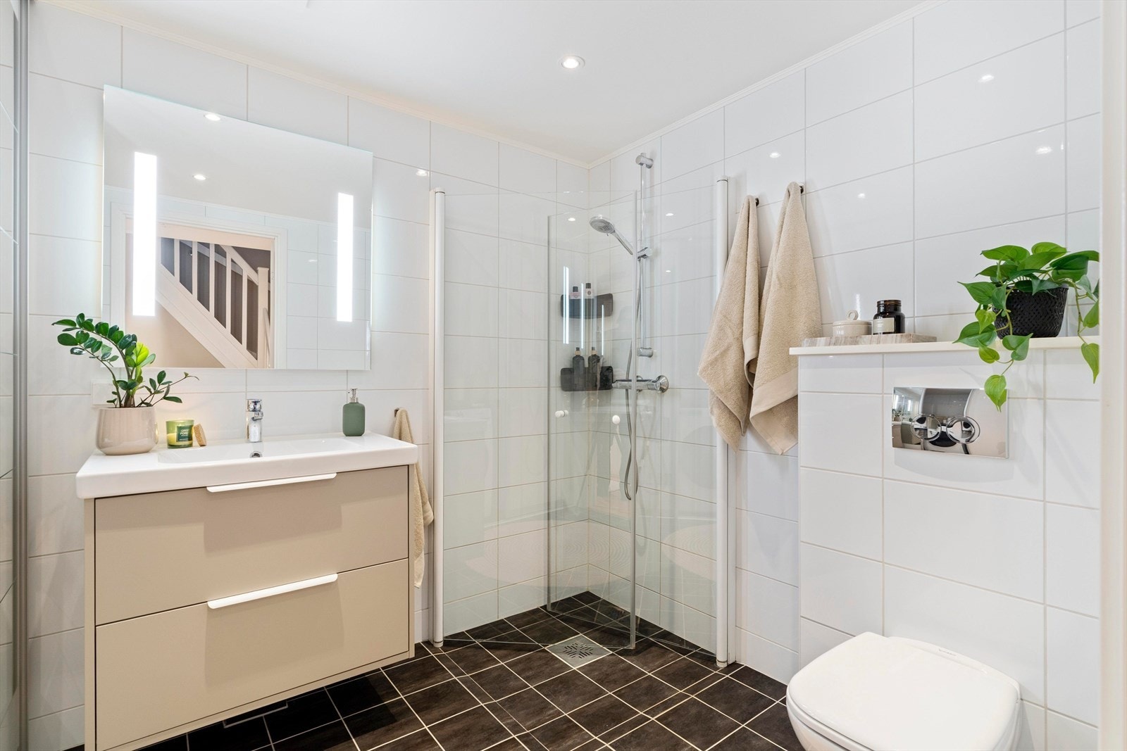 Badet fremstår som moderne og helhetlig med flislagte gulv og vegger. Rommet er innredet med vegghengt toalett, dusj med glassvegger, servant med underskap og speil med belysning. Galleribilde