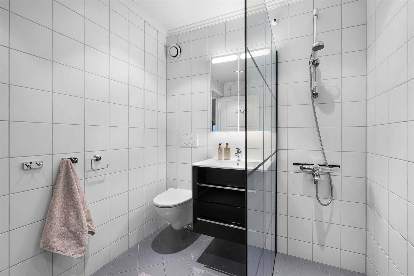 Boligens bad er helfliset og innredet med vegghengt WC, dusjhjørne, servant med underskap, speil og varmekabler i gulvet. Galleribilde