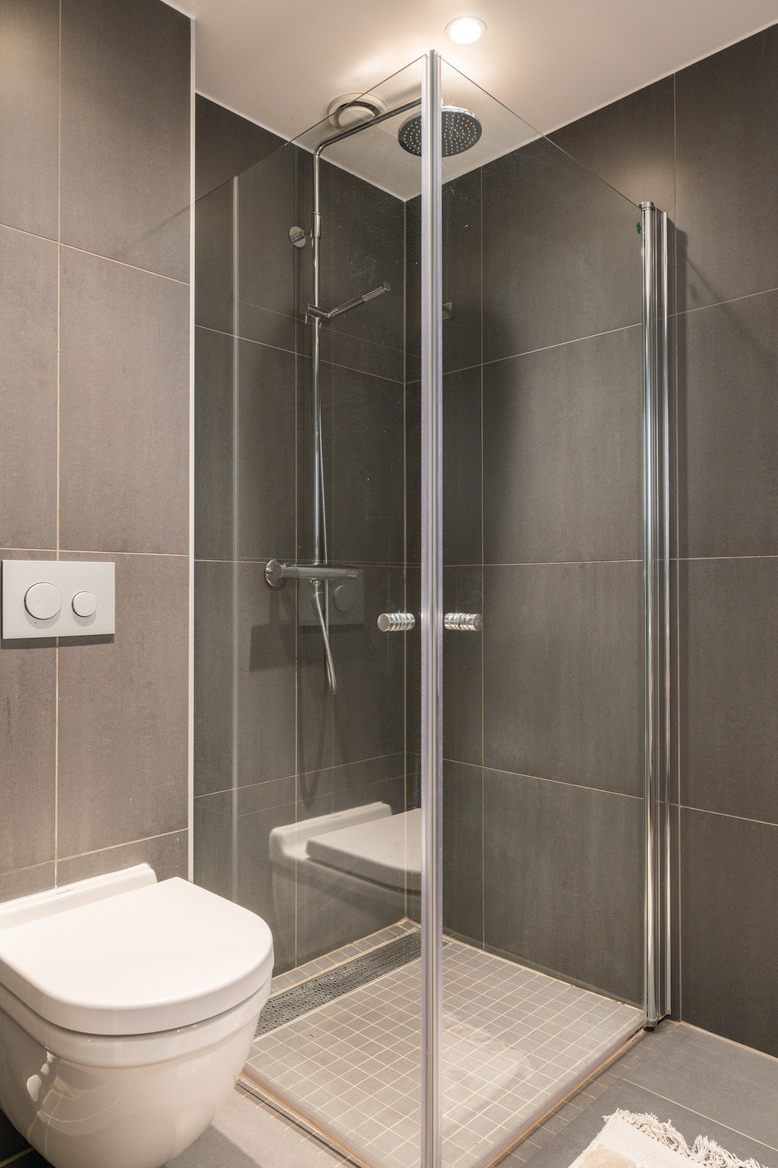 Dusjhjørne med mosaikkfliser på gulv, innfellbare glassdører og armatur med både hånd- og regnfallsdusj. Galleribilde