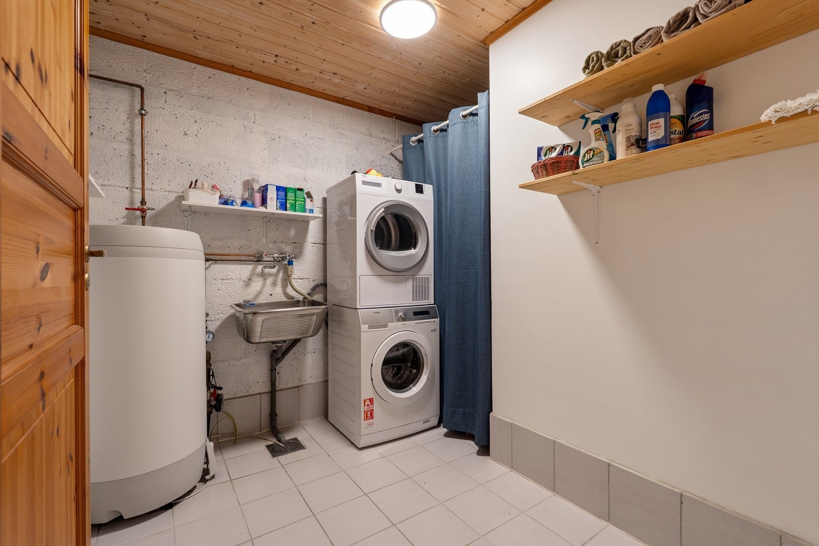 Separat vaskerom i underetasje med fliser på gulv og malte overflater på vegg. Skyllekar, varmtvannsbereder og opplegg for vaskemaskin. Det er naturlig ventilasjon. Galleribilde