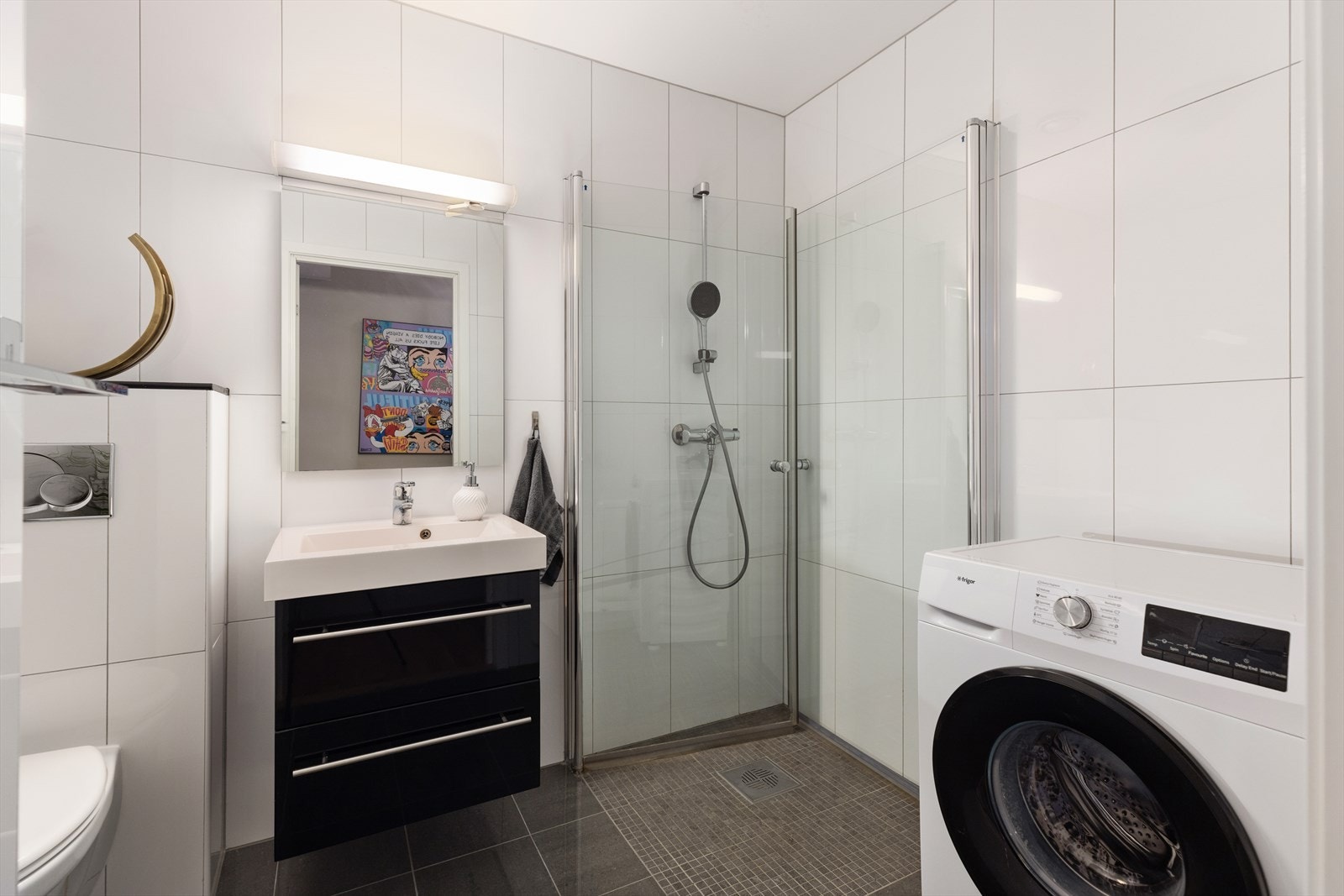 Pent helfliset bad utstyrt med vegghengt toalett, servant i skap, dusjsone med innfellbare glassdører, samt opplegg for vaskemaskin Galleribilde