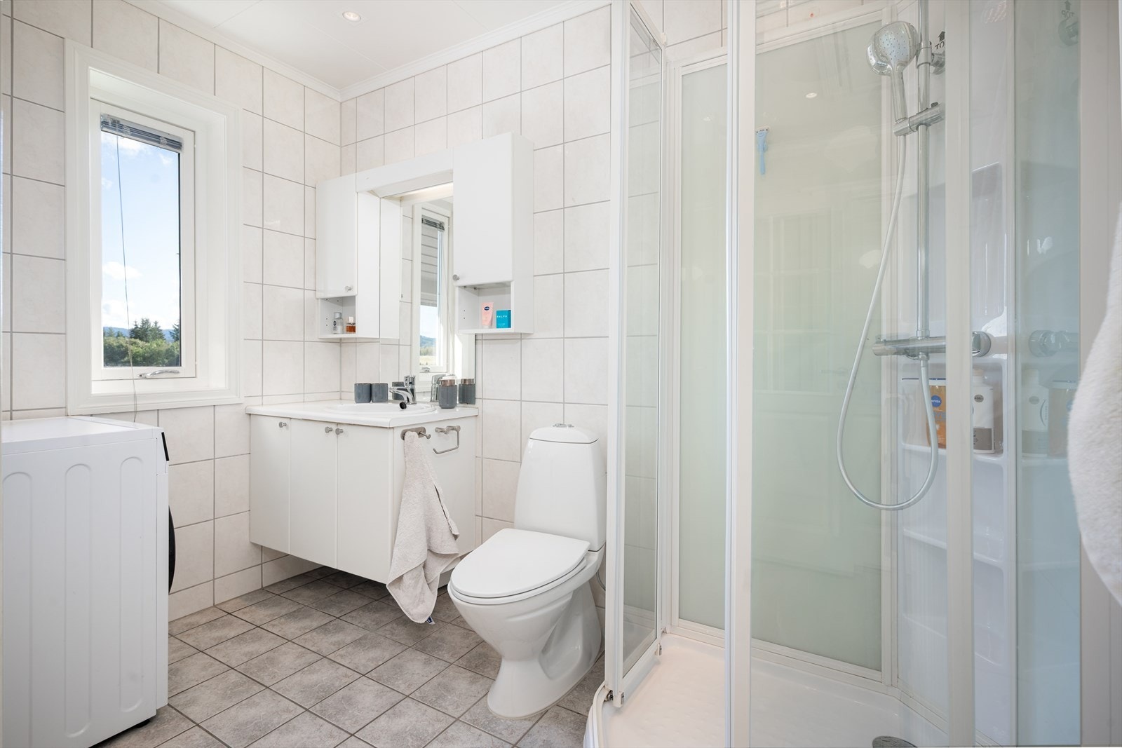 Badet er flislagt og utstyrt med varmekabler i gulv, hvit servantinnredning med god plass til hygieneartikler og dusjkabinett. Galleribilde