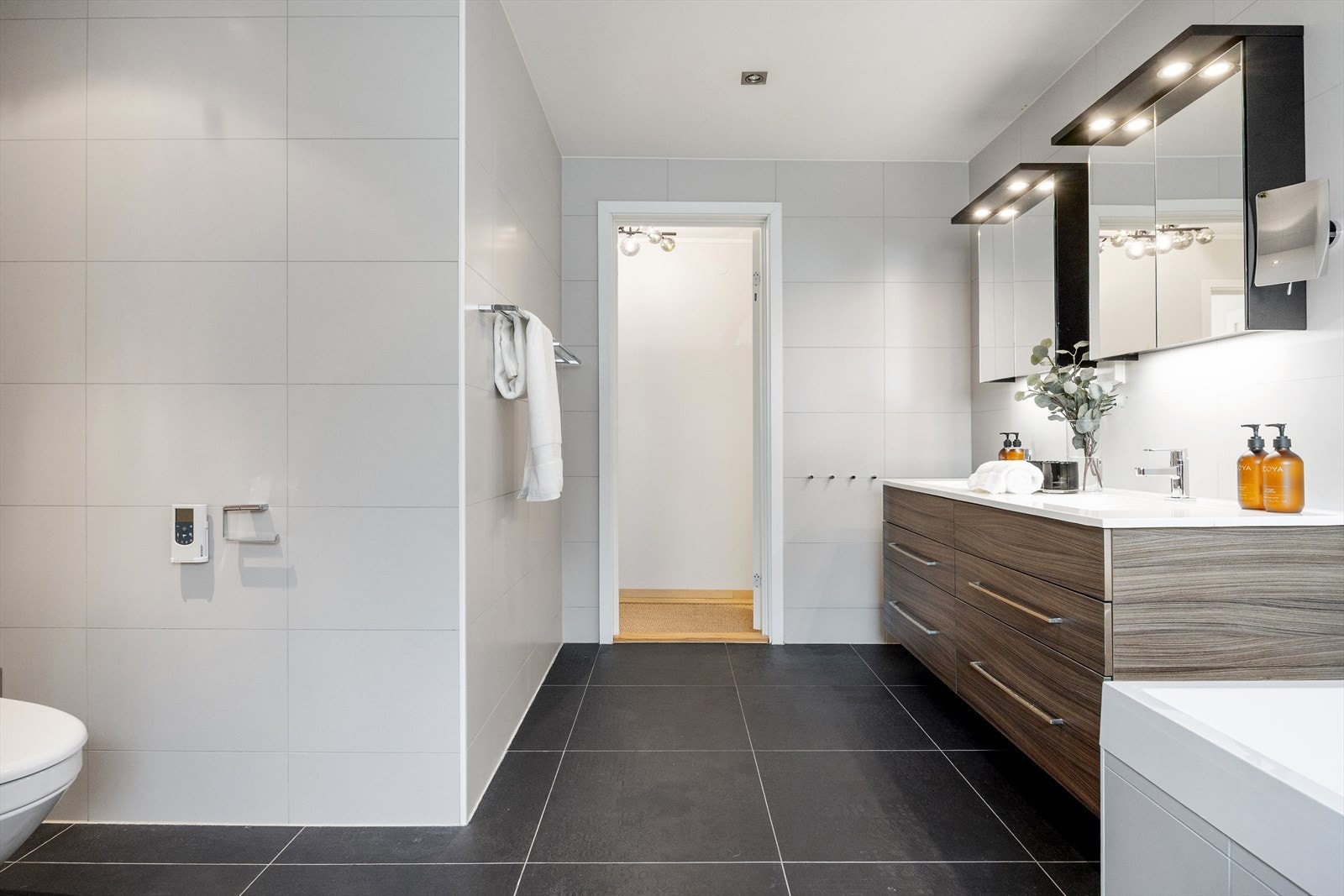 Rommet er innredet med veggmontert spyletoalett, badekar, dusjnisje og baderomsinnredning med to servanter, ettgreps blandebatteri, skuffer og vegghengte speilskap med belysning. Galleribilde