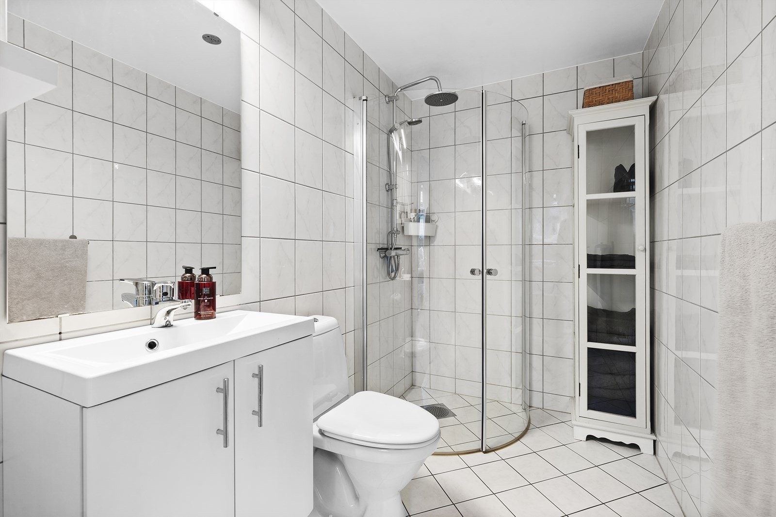 Lyst og helfliset baderom med varmekabler i gulv. Badet er utstyrt med servant over skapinnredning, speil med overliggende belysning, toalett og dusjhjørne med innfellbare glassvegger Galleribilde