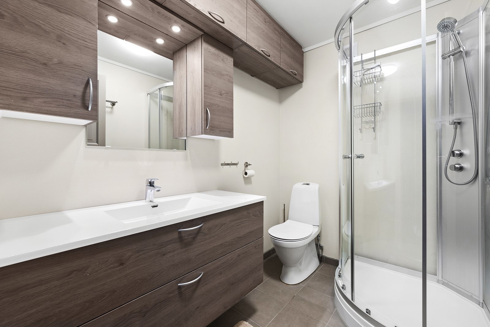 Lyst baderom med flislagt gulv med varmekabler. Badet er innredet med servant over skuffeinnredning, vegghengt skap, toalett og dusjkabinett Galleribilde