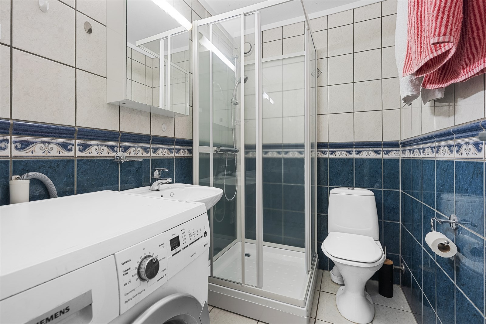 Flislagt baderom med varmekabler i gulv. Badet er utstyrt med servant, speilskap med overliggende belysning, toalett, dusjkabinett fra desember 2025 og opplegg til vaskemaskin Galleribilde