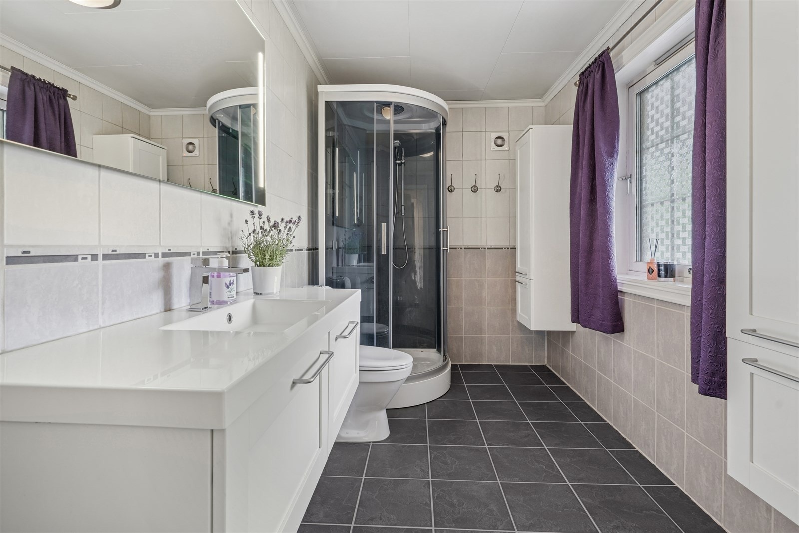 Badet er utstyrt med heldekkende servant, speil med lys, to høyskap, dusjkabinett og gulvmontert toalett. Galleribilde