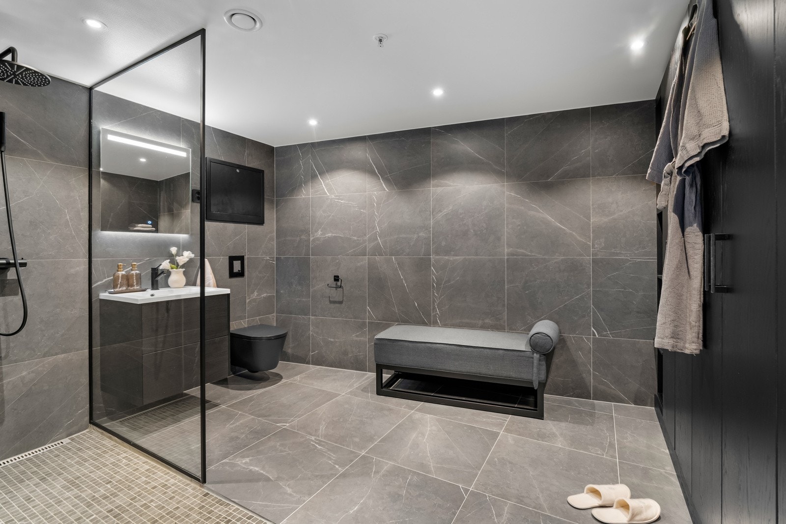 Badet har et strøkent uttrykk med store, sorte vegg- og gulvfliser i marmormønster. Langs den ene veggen er det satt inn sorte, spesialtilpassede garderobeskap i heltre, levert av Drømmekjøkkenet. Galleribilde