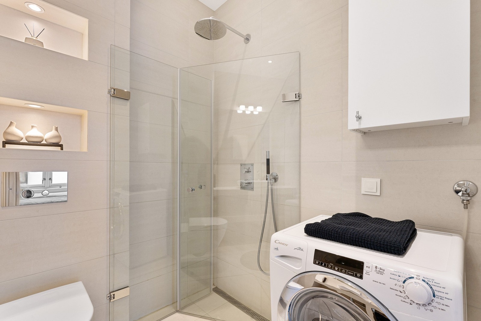 Badet er innredet med møbelservant på fliselagt benkeplate, veggmontert toalett og dusjløsning med glassvegger. Videre er det speil med integrert belysning og opplegg for vaskemaskin. Galleribilde