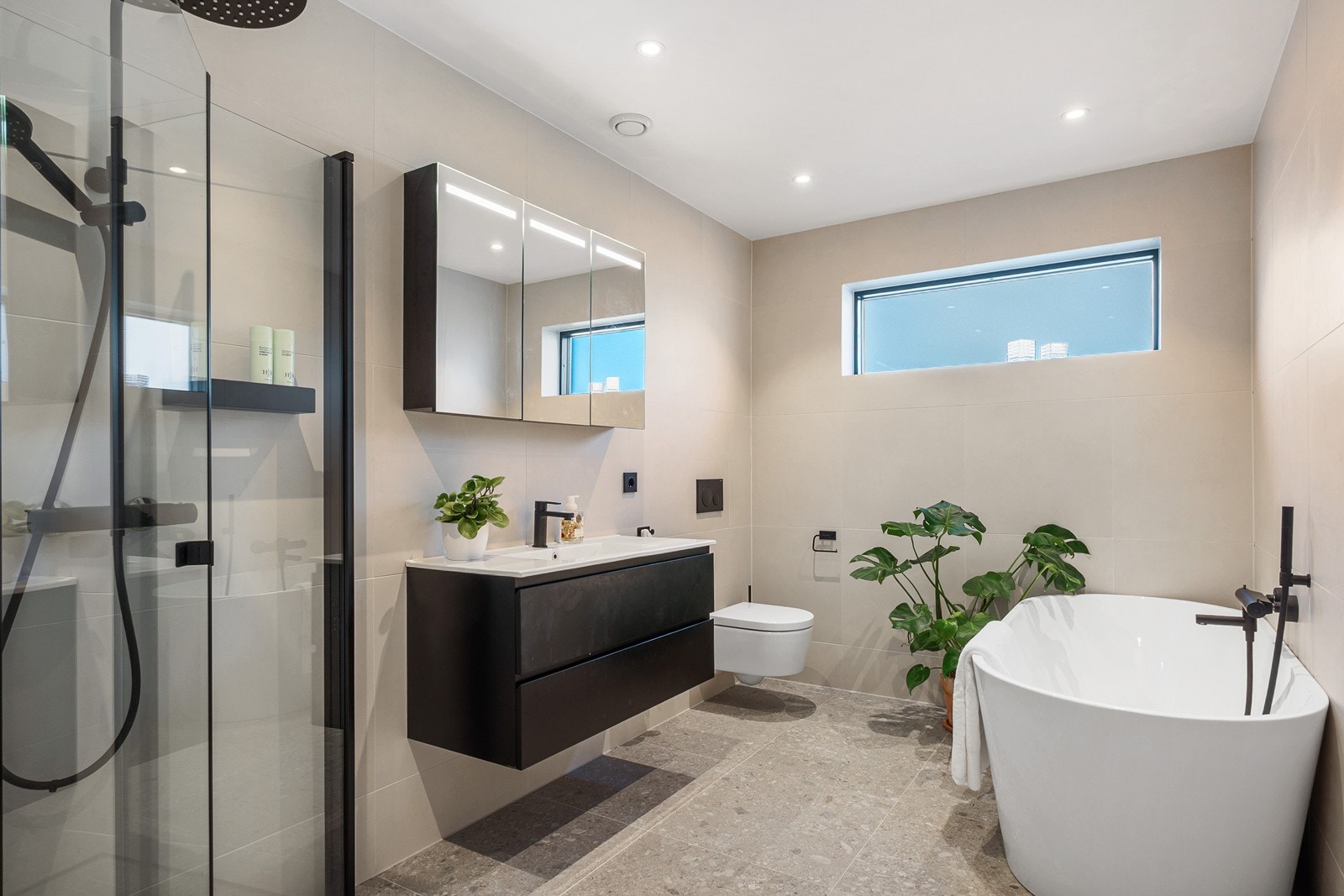 Badet i første etasje er innredet med lyse 60x60 fliser, sorte detaljer og armaturer. Badet har servant med skuffeseksjon, speil og vegghengt toalett, samt et dusjhjørne med innfellbare glassdører, hånd- og takdusj - i tillegg til badekar. Galleribilde