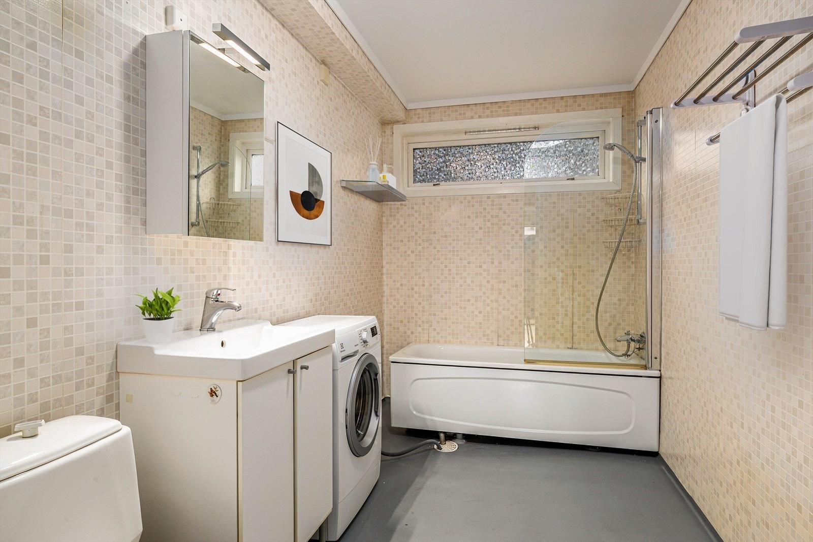 Eldre bad/vaskerom (TG 3) med belegg på gulv og elektriske varmekabler, samt vegger med våtromstapet (ikke målt fukt ved badekaret). Taket har malte plater. Rommet er innredet med badekar, servantskap, klosett, hylle og opplegg til vaskemaskin. Galleribilde