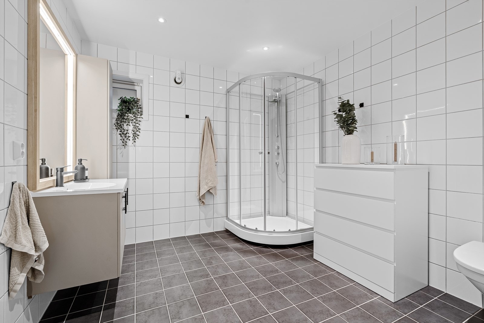 Badet i leilighetens hovedetasje er helfliset med varme i gulvet og downlights i himling. Det inneholder et praktisk dusjkabinett, vegghengt toalett, samt baderomsinnredning med eget designet eikerammet speil med god sminkebelysning. Galleribilde