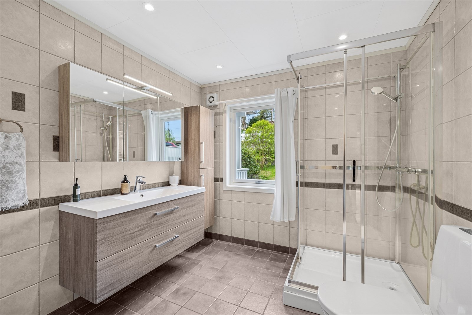 Flislagt bad/wc/vaskerom med varmekabler i gulv og platekledd himling med integrert belysning. Rommet er utstyrt med hel nedfelt servant, klosett, dusjkabinett, opplegg for vaskemaskin og baderomsinnredning med servantskap, speilskap og to høyskap. Galleribilde