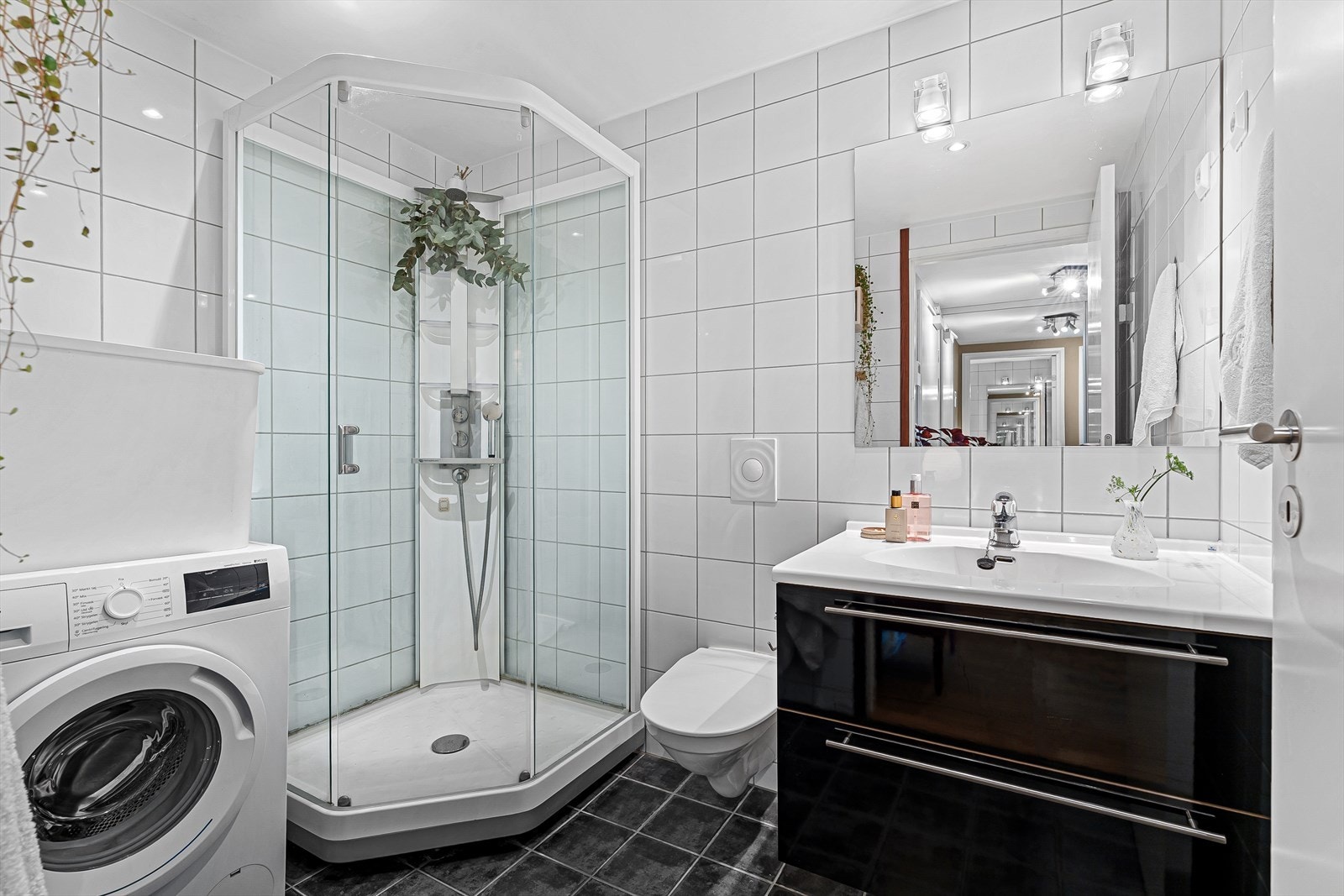 Badet er helfliset og inneholder et praktisk dusjkabinett, vegghengt toalett, baderomsinnredning med speil og belysning over, samt opplegg for vaskemaskin. Galleribilde