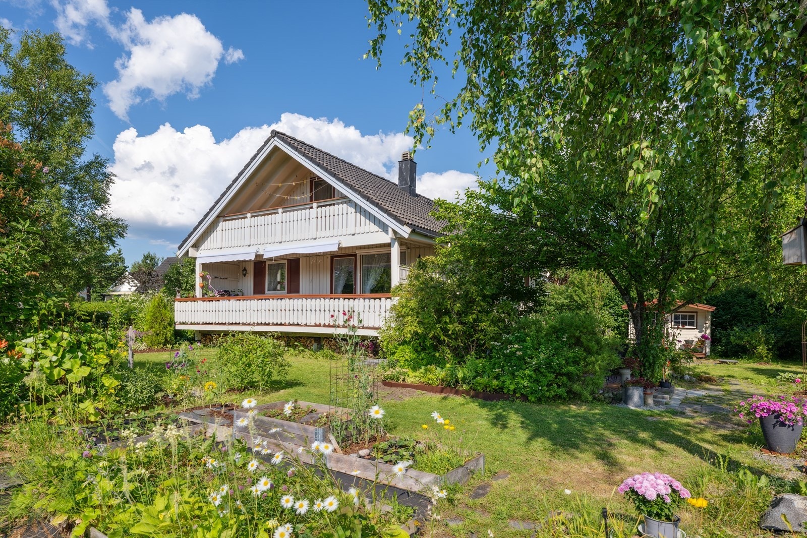 Solrik og vakker hage med plen og beplantning. Uthus til høyre i bildet. Galleribilde