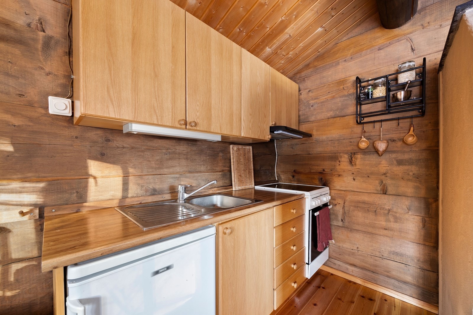 Kjøkkenet er utstyrt med komfyr med kokeplater, ventilator, vask og kjøleskap. Innredningen har slette dører og skuffefronter i eik, samt en benkeplate i laminat med benkebeslag i stål. Galleribilde