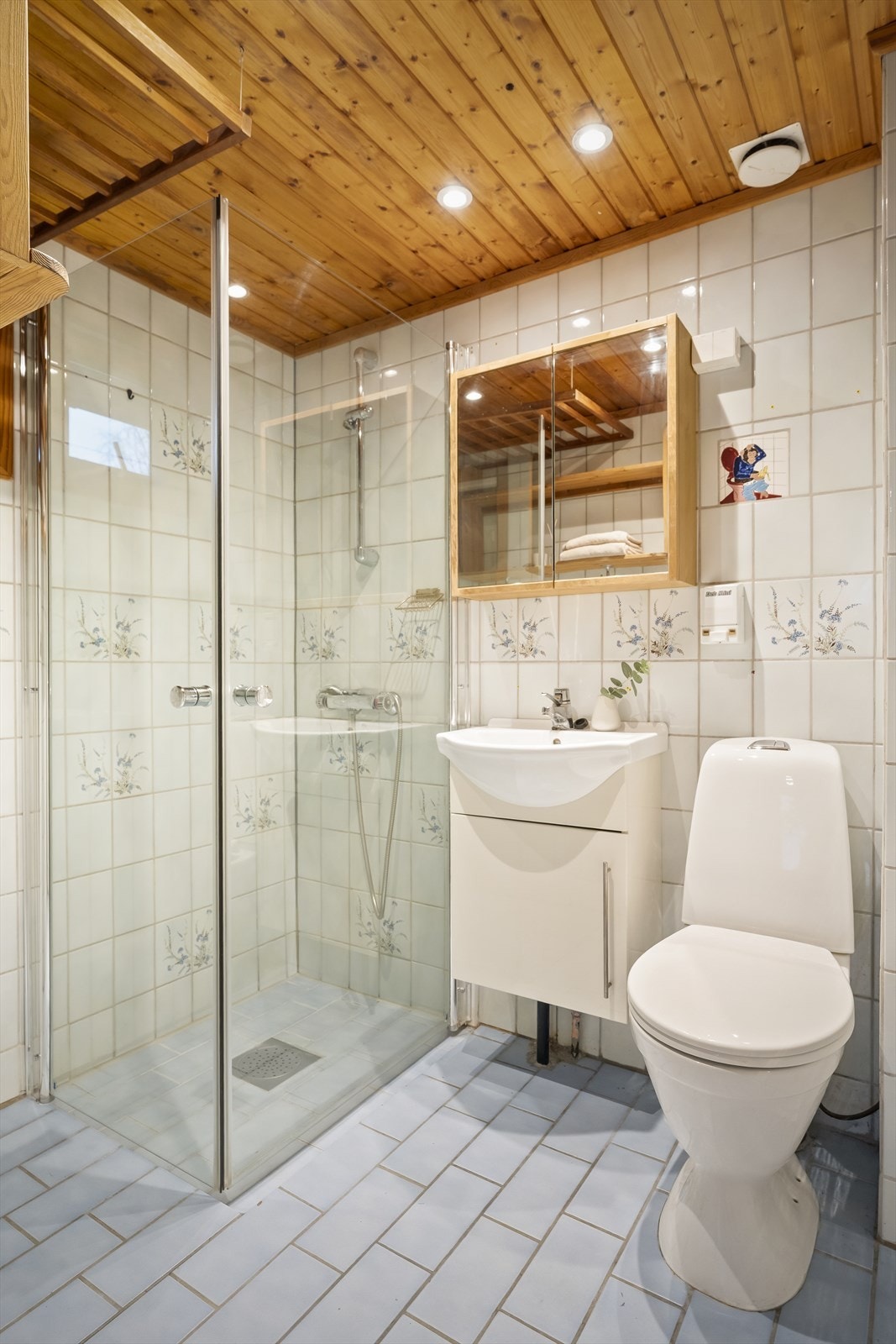 Bad i første etasje med fliser på gulv og vegger. Rommet er utstyrt med et dusjhjørne med svingbare glassdører, servant med skap og toalett. Galleribilde