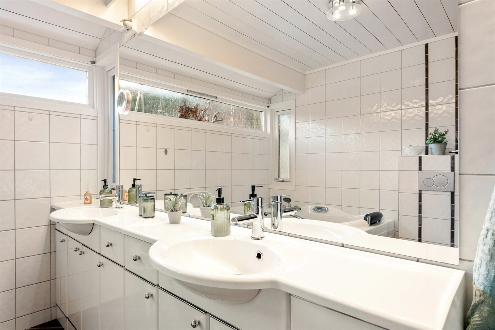 Stor og innholdsrik baderomsinnredning med hvite glatte fronter og doble servanter med ett-greps armatur. Stort speil med overlys over servanter. Galleribilde