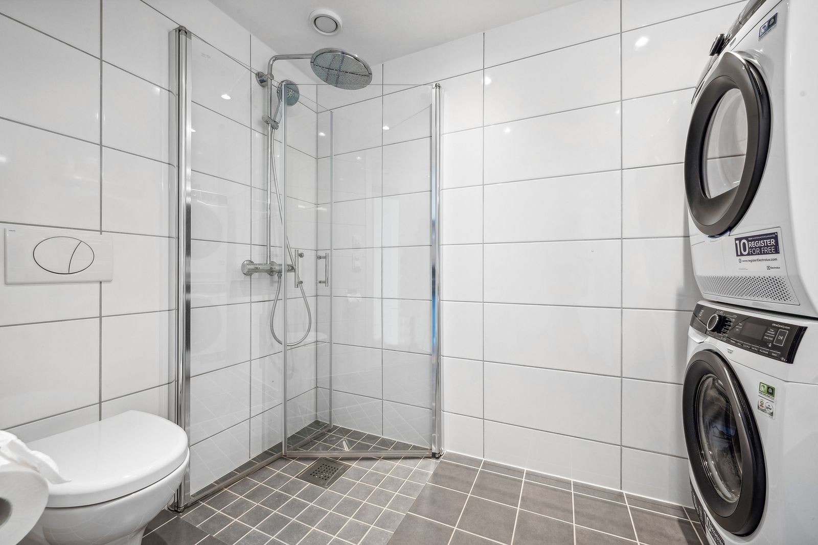 Pent flislagt bad innredet med dusjdører i glass, og opplegg for vaskemaskin og tørketrommel. Galleribilde