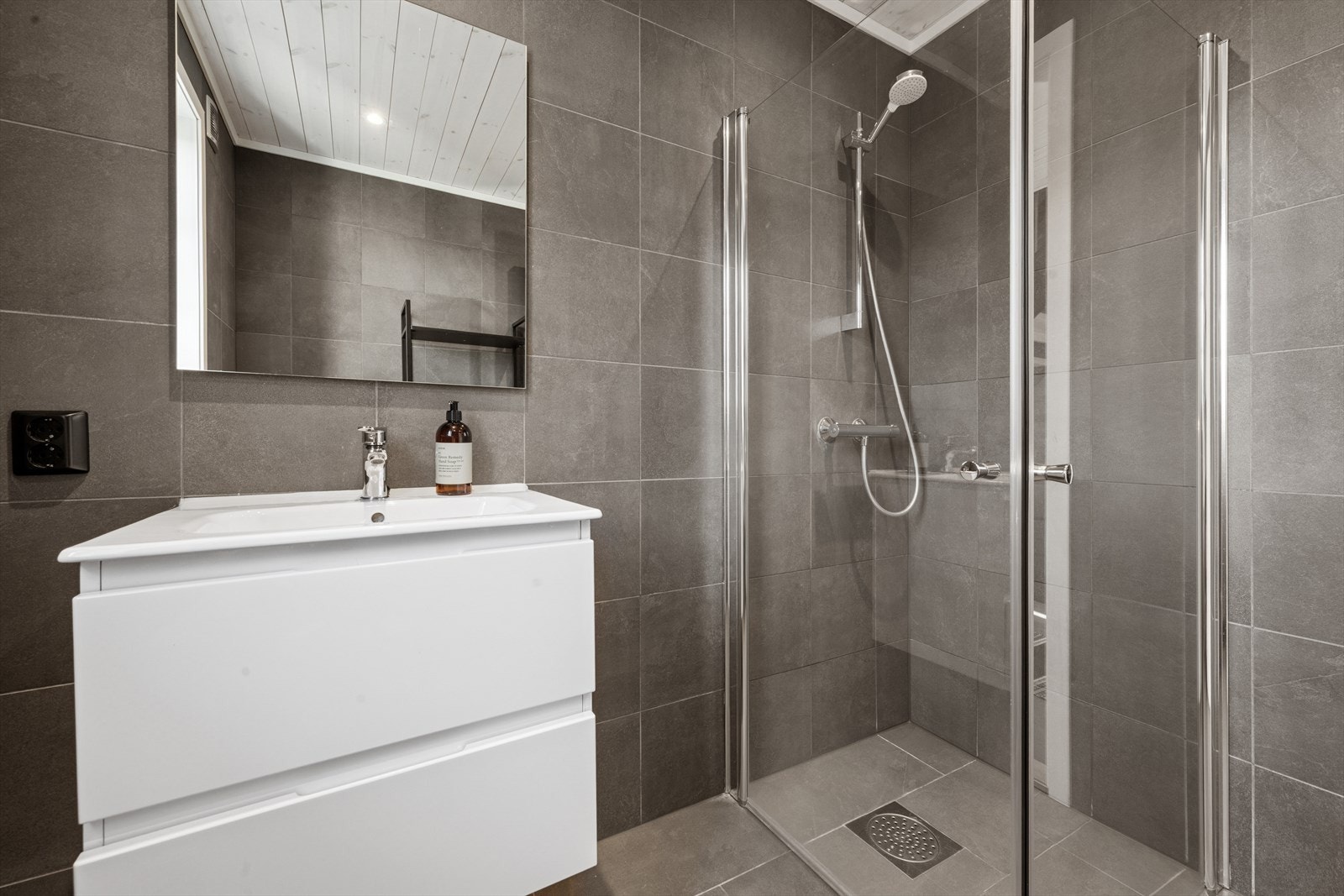 Badet i første etasje er stilfullt innredet og har fliser i lekre, mørke fargetoner. Her finner du dusj med glassdører, moderne baderomsinnredning med heldekkende servant, skuffer og speil. Galleribilde