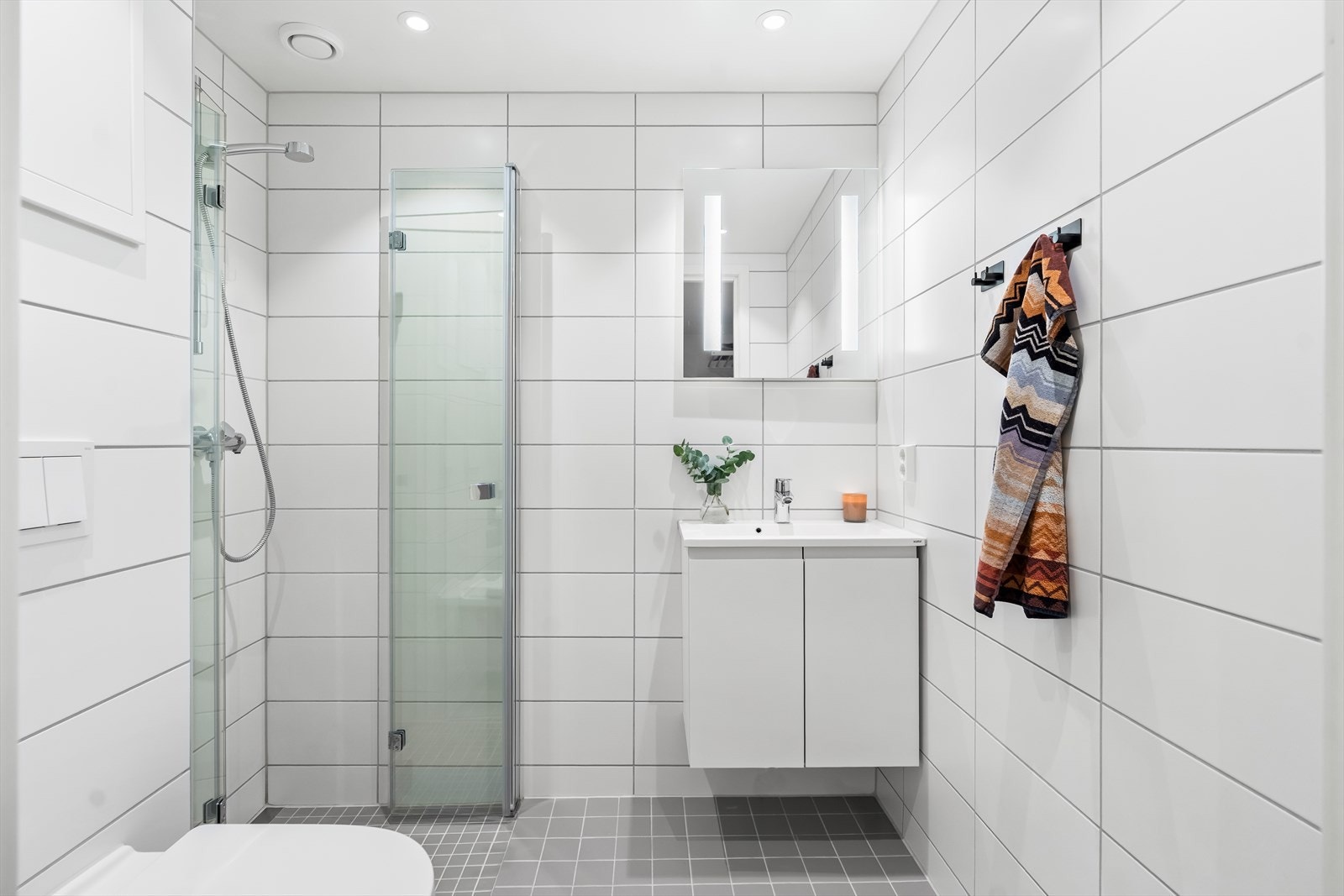 Fra gangen er det inngang til et flislagt baderom. Rommet har dusjhjørne med dører i herdet glass, veggfestet klosett med innebygget sisterne samt servant. Galleribilde