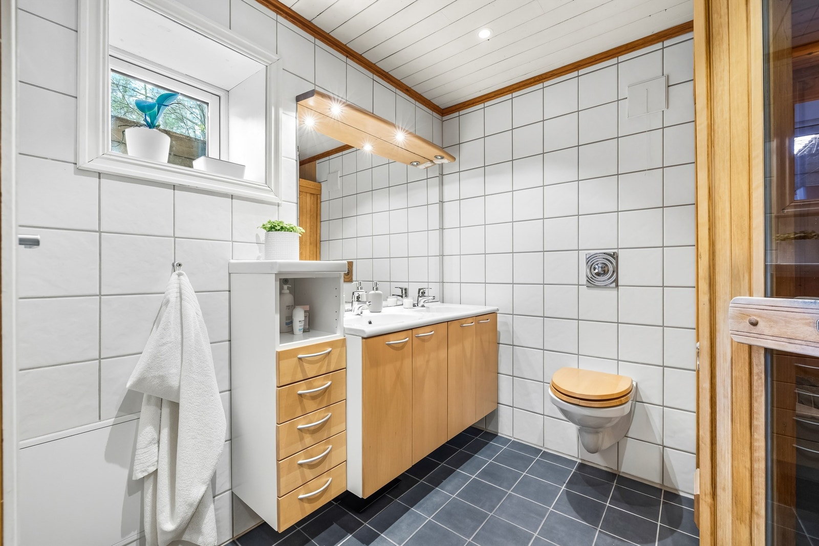 Badet er innredet med dusjkabinett med glassdør og dusjarmatur, vegghengt toalett og vegghengt servantskap med to heldekkende servanter. Galleribilde