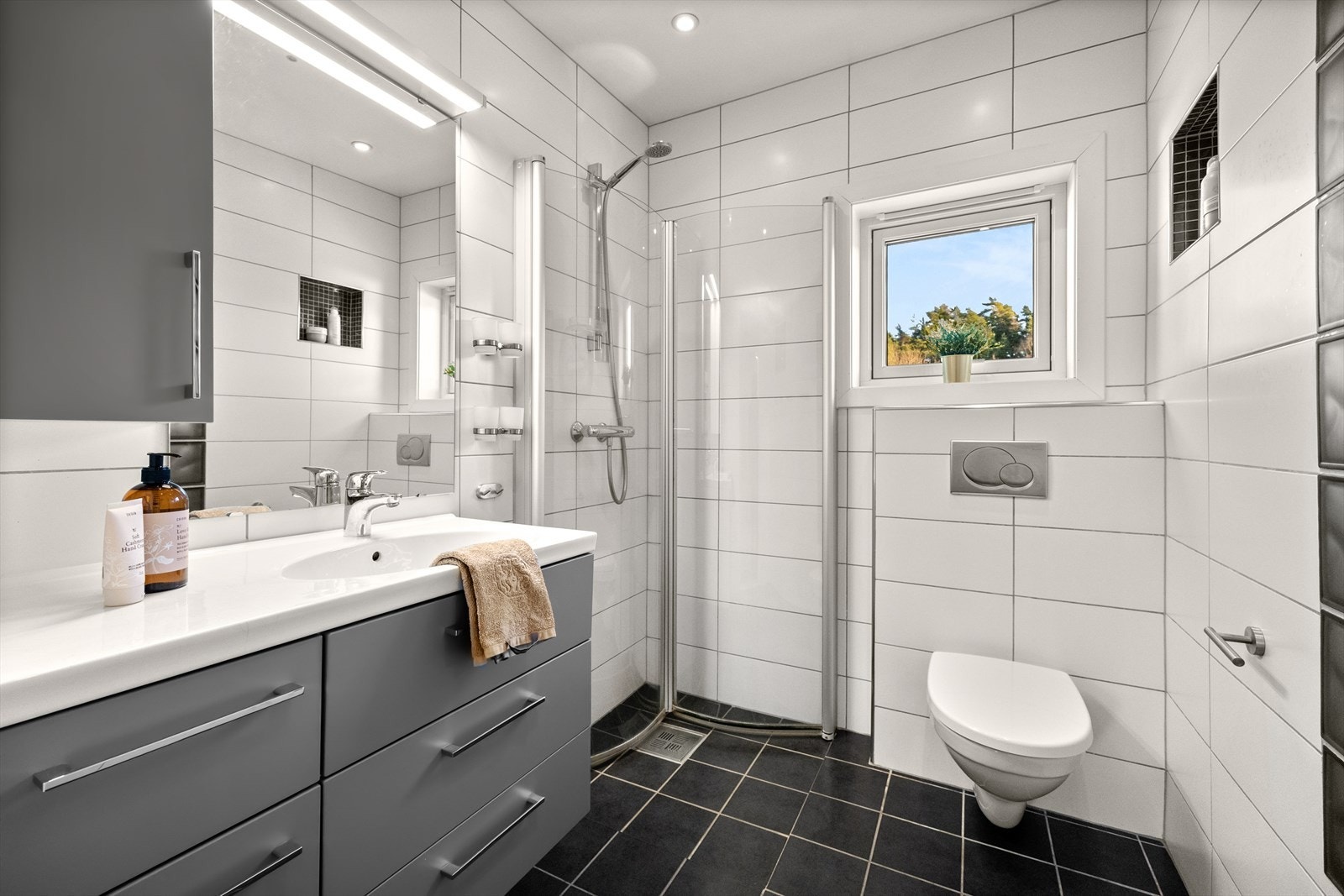 Rommet er innredet med baderomsinnredning med heldekkende servant, speil, veggfestet klosett og dusj med dører i herdet glass. Galleribilde