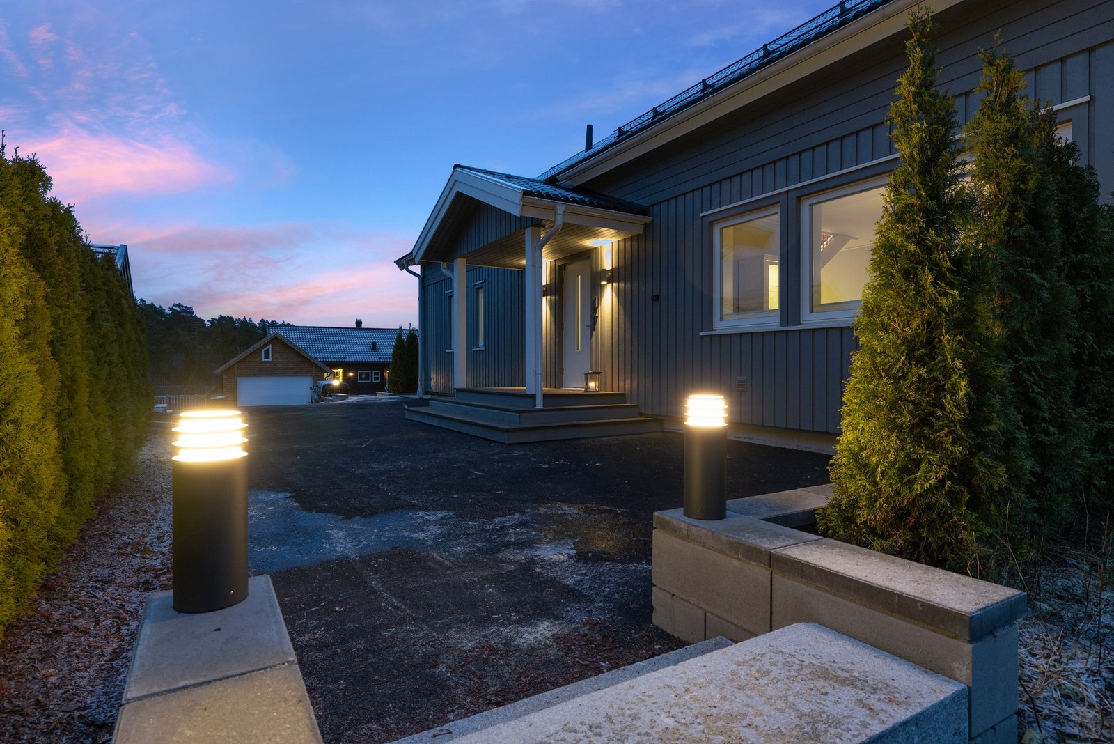 Fasade i front med fin lyssettning. Eiendommen er stort sett omkranset med vintergrønn hekk. Galleribilde