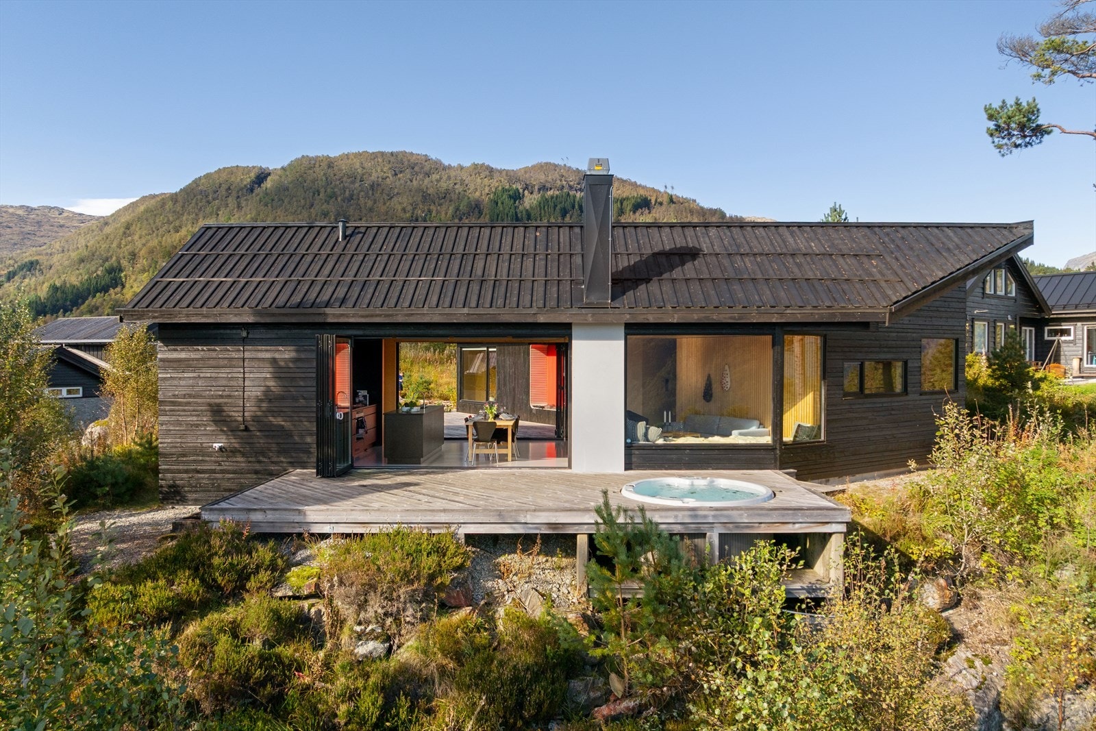 Ytre materialbruk er mørkbeiset royalbehandlet kledning. Det er foldedører i sort aluminium på hver side av stuen/kjøkkenet. Galleribilde