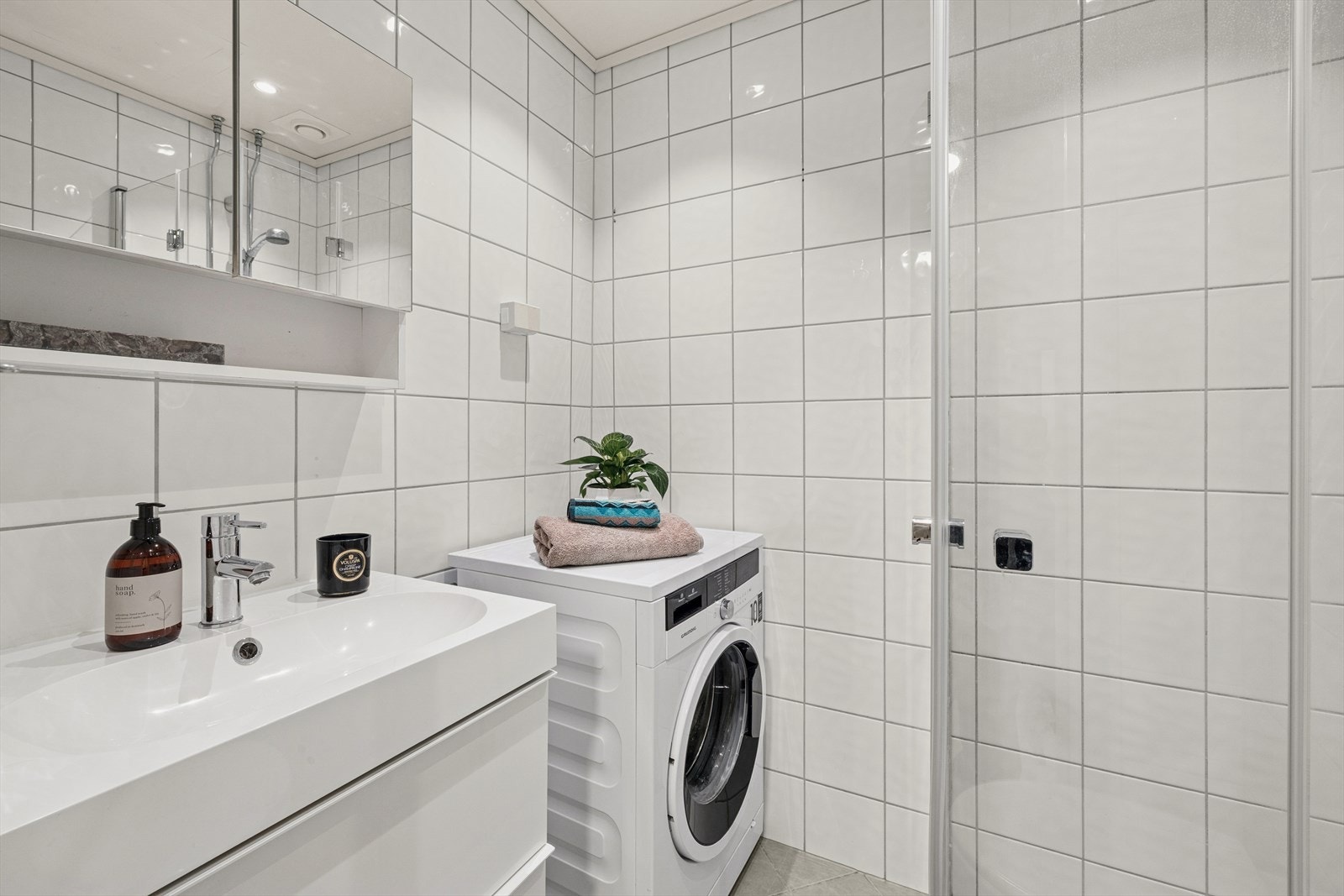 Rommet er innredet med en baderomsinnredning med benkeskap, veggskap med speil, servant, veggfestet klosett med innebygget sisterne og dusj. Galleribilde