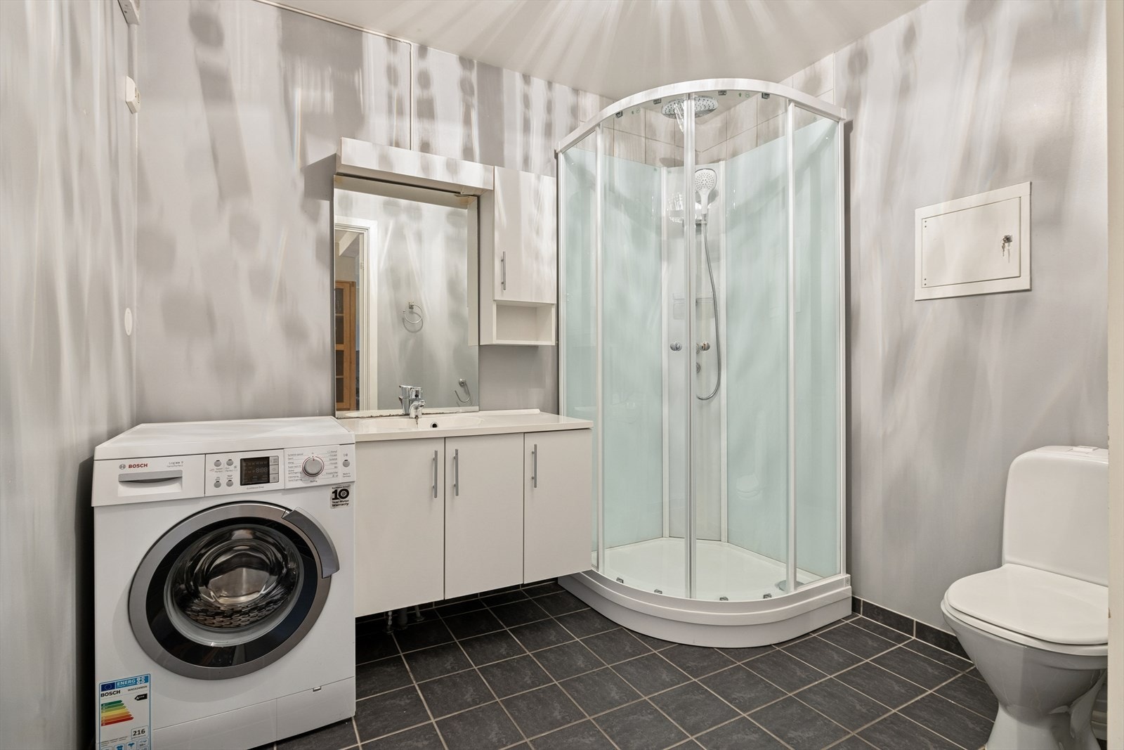 Baderom med flislagt gulv, dusjkabinett, toalett, opplegg til vaskemaskin og servant med oppbevaringsplass. Galleribilde