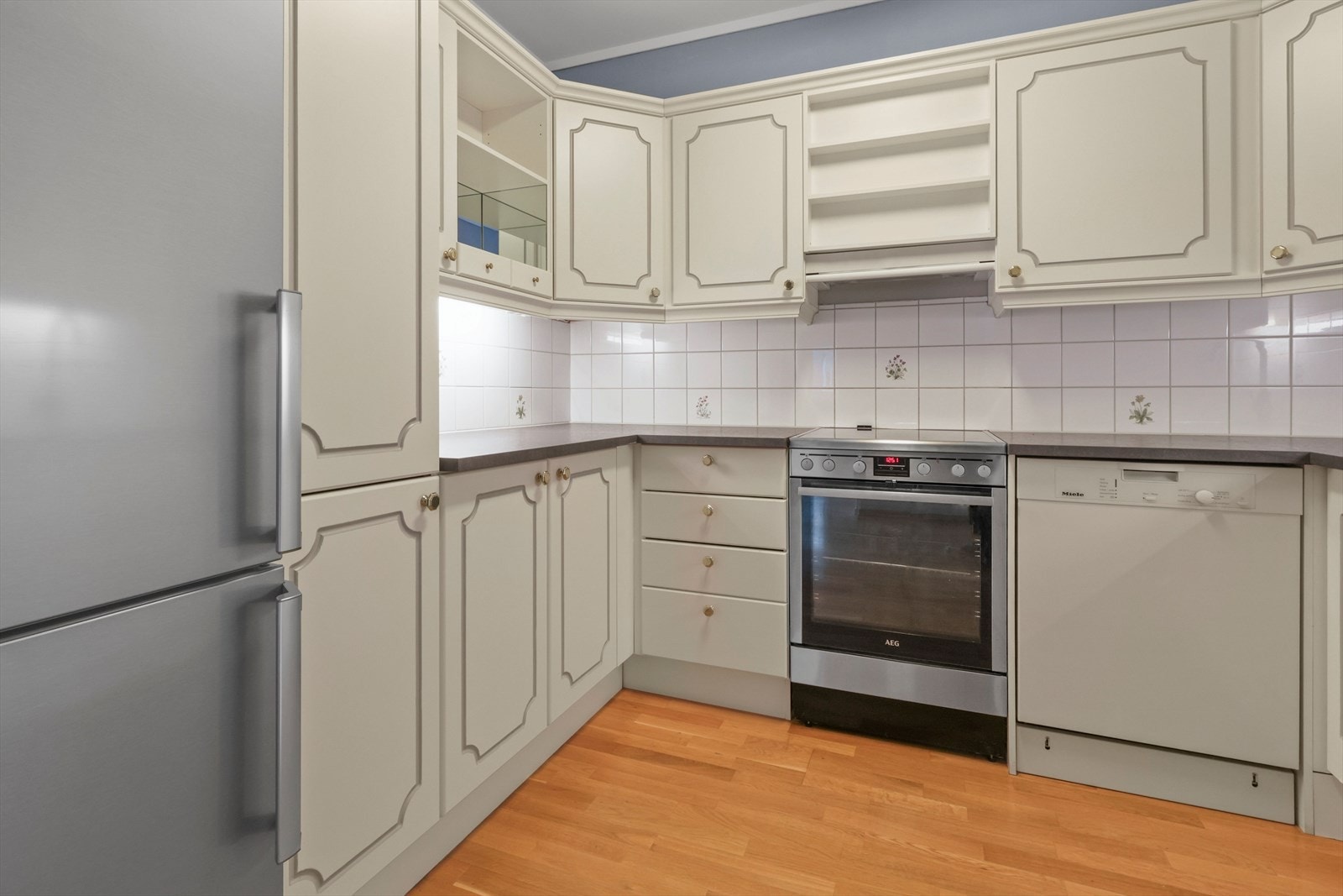 Lys profilert kjøkkeninnredning med komfyr, oppvaskmaskin og kombinert kjøleskap/fryser. Galleribilde