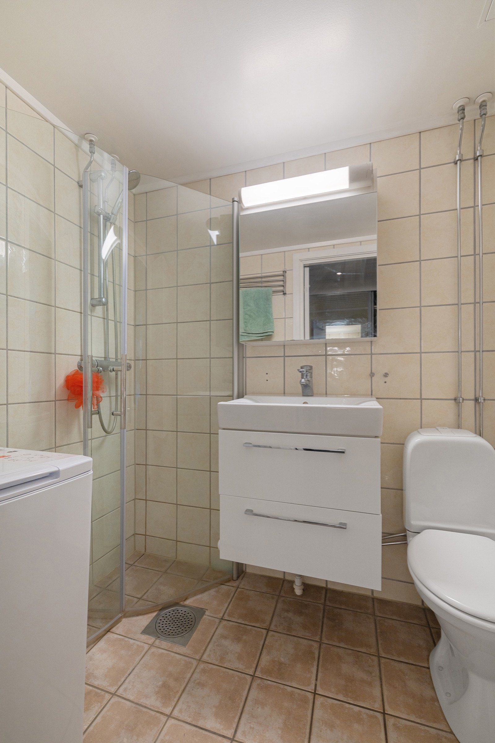 Rommet er innredet med gulvmontert toalett, nedsenket servant med tilhørende innredning og speil, dusjnisje med glassdører samt opplegg for vaskemaskin. Galleribilde