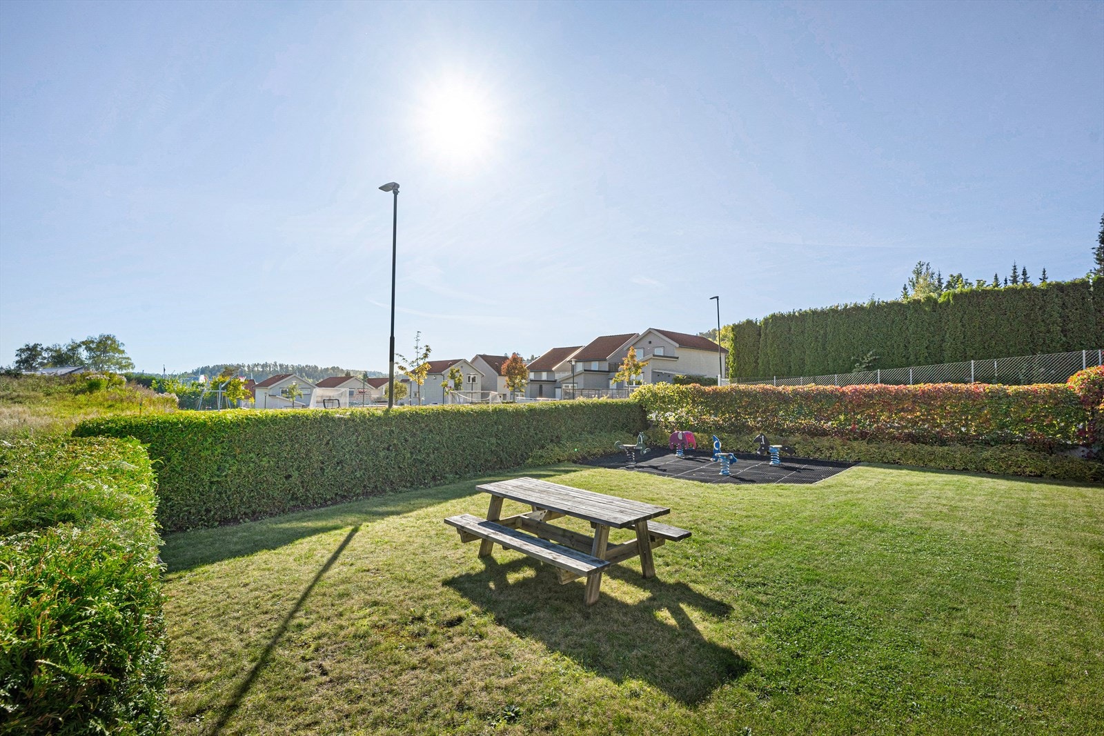 Fellesområde med benk, bord og en mindre lekeplass. Over veien er det en større lekeplass og fotballbane. Galleribilde