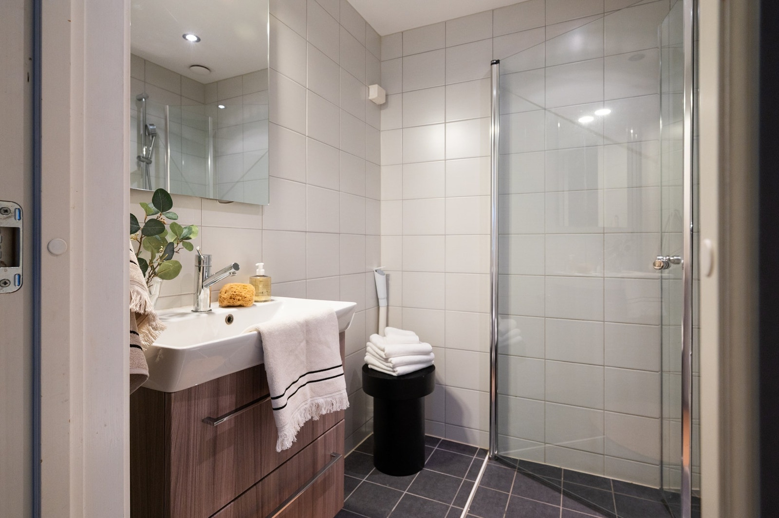 Flislagt bad med mørke gulvfliser, lyse veggfliser og dusjhjørne med innfellbare glassdører. Galleribilde