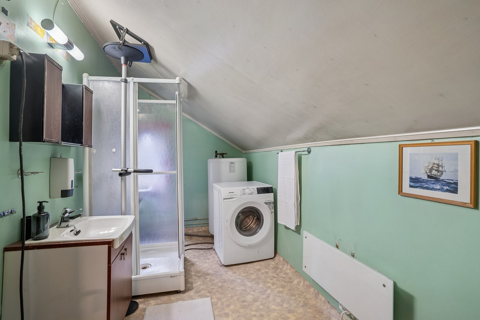 Baderommet er utstyrt med servantinnredning, dusjkabinett, varmtvannsbereder og wc på sokkel. Det er malte plater i taket, tapet på veggene og belegg på gulvet. Overflatene fremstår med elde/slitasje, og må renoveres i sin helhet. Galleribilde