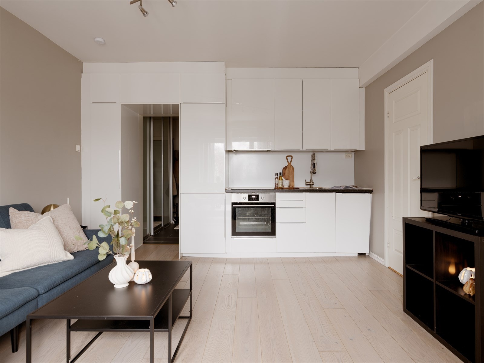 Leiligheten har en åpen stue-kjøkkenløsning med smart kjøkkeninnredning i hvite glatte fronter som rammer inn inngangspartiet. Galleribilde