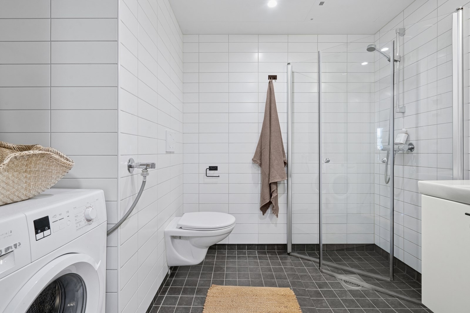 Romslig og flislagt baderom med tidløs innredning bestående av servant med speil, vegghengt toalett og dusjhjørne. Det er også opplegg for vaskemaskin. Plass til tørketrommel. Varmekabler i gulv. Galleribilde