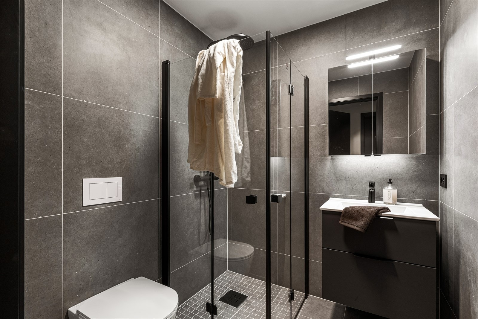 Delikat helfliset bad i 1. etasje med varmekabler i gulv og downlights i hiling. Badet er innredet med vask i seksjon, vegghengt toalett og dusjhjørne. Galleribilde